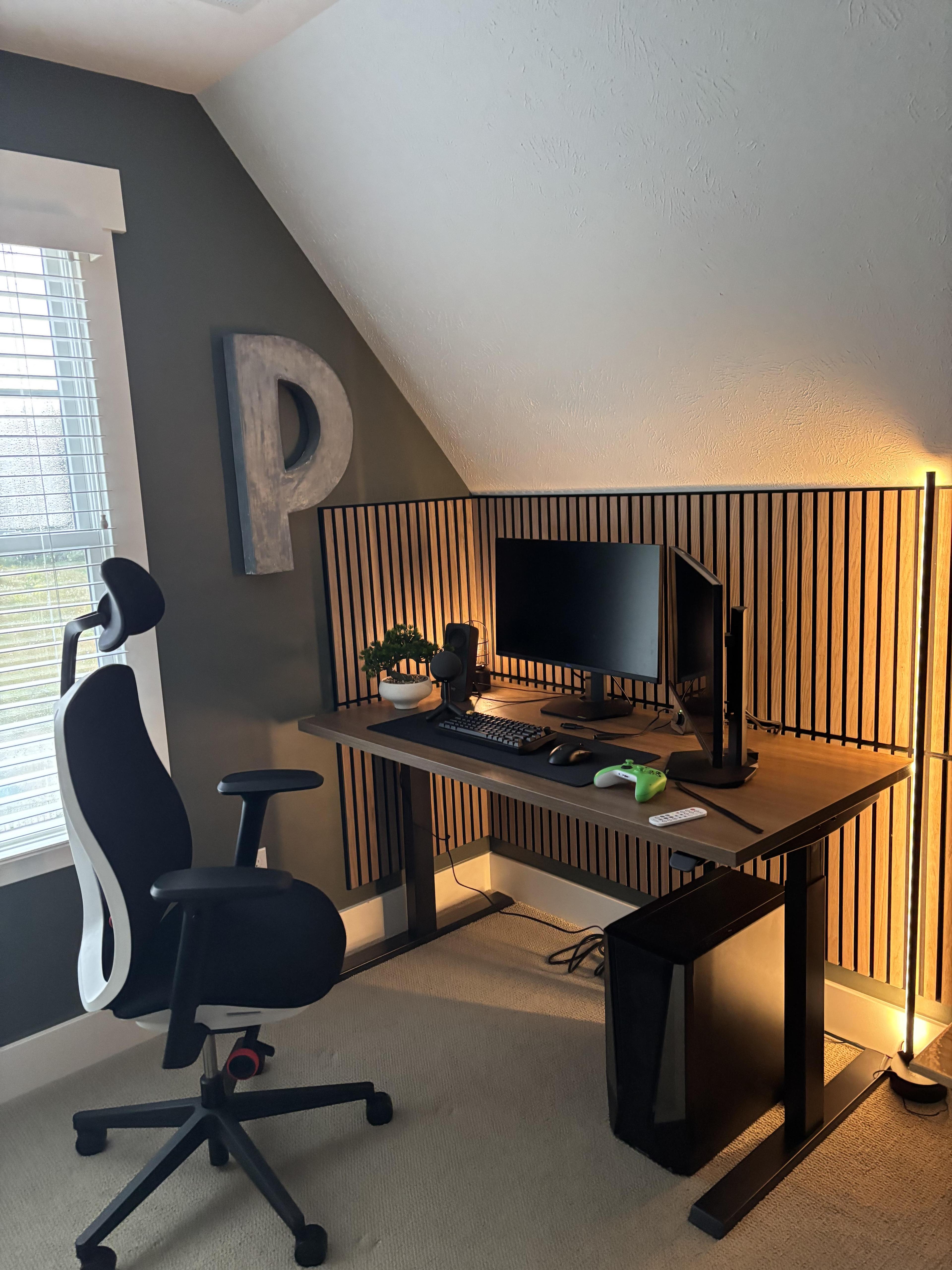 A corner desk under a sloped ceiling, with wood slat panels over two walls, two monitors, a black chair and a tall warm light bar on the right.