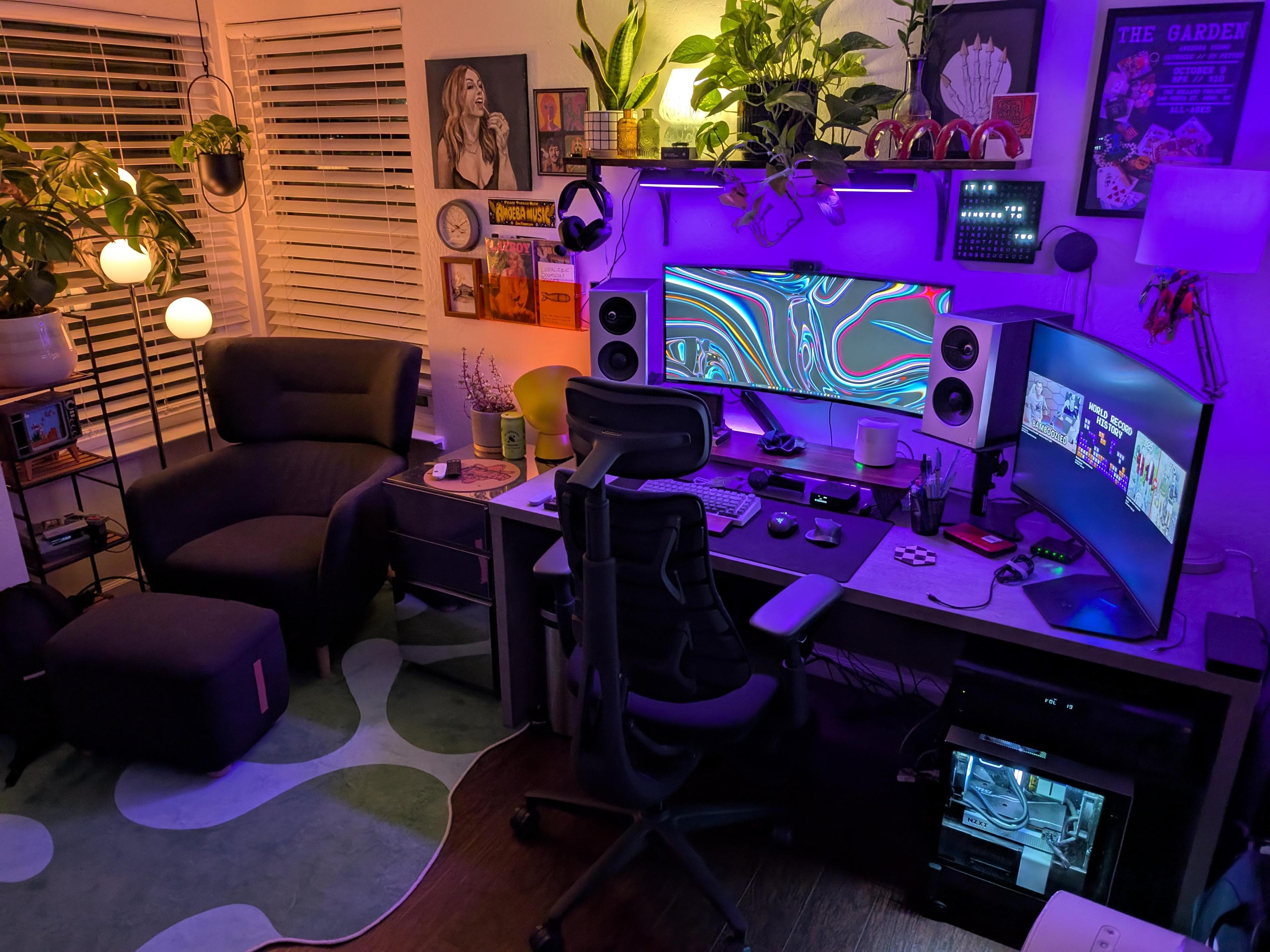 A home office lit purple. An ultrawide screen on a desk with a speaker either side, a second screen turned side on at the right, a dark armchair and footstool by a shuttered window, plants on a stand, and a wall of small framed art.