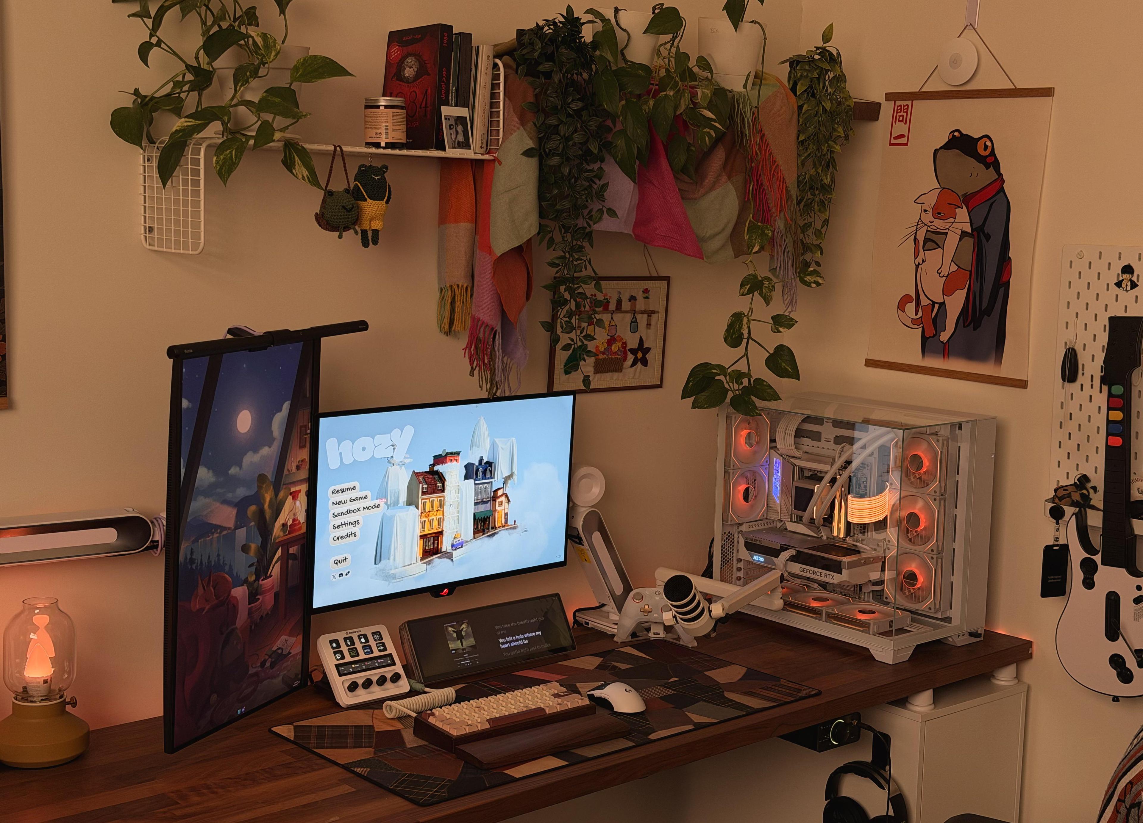 A warm lit wooden desk under a shelf of trailing plants and hanging scarves, with a vertical screen, a main monitor, a lyric screen, a custom keyboard and a white water cooled PC on the right.