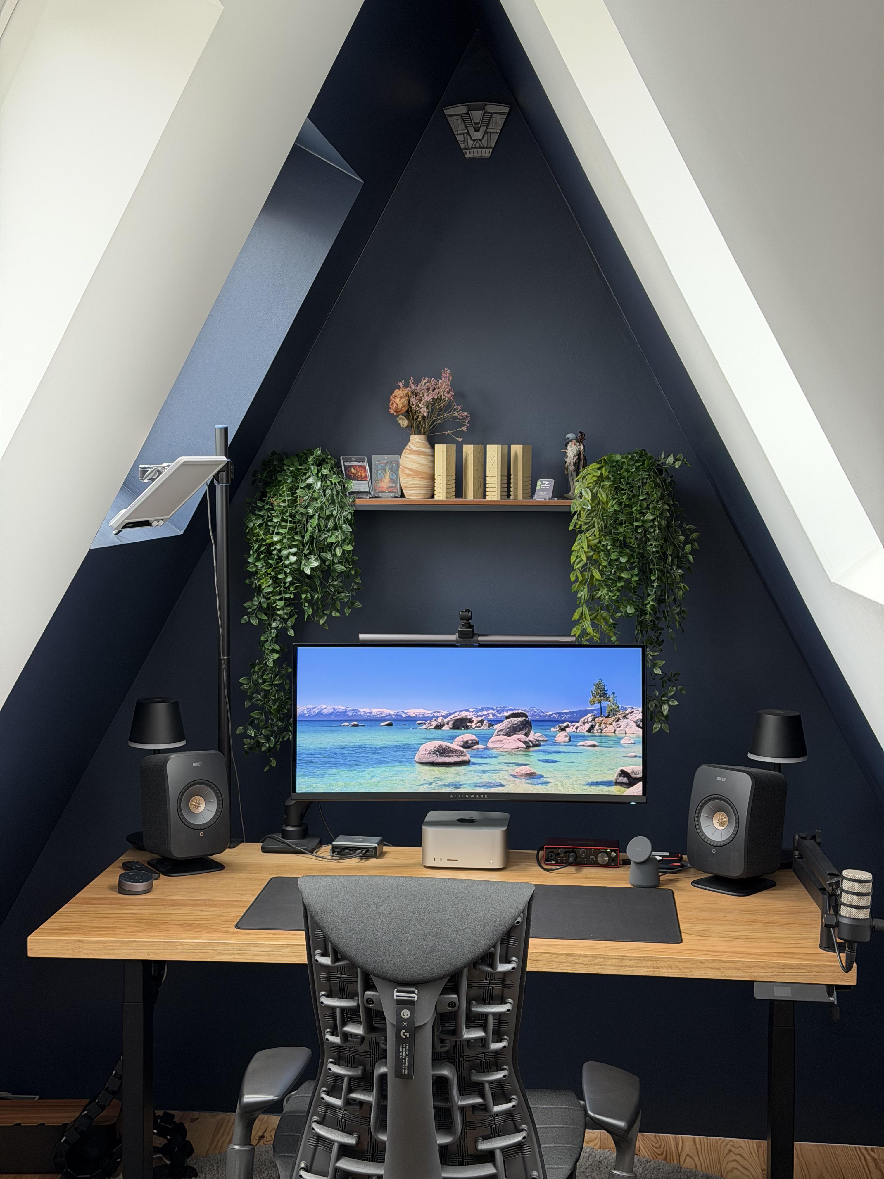 A desk built into a triangular attic nook painted dark blue. An ultrawide screen on an arm, a small computer on the wooden top, a speaker with a lamp behind it either side, a shelf of ornaments and dried flowers, and a trailing plant at each end of the shelf.