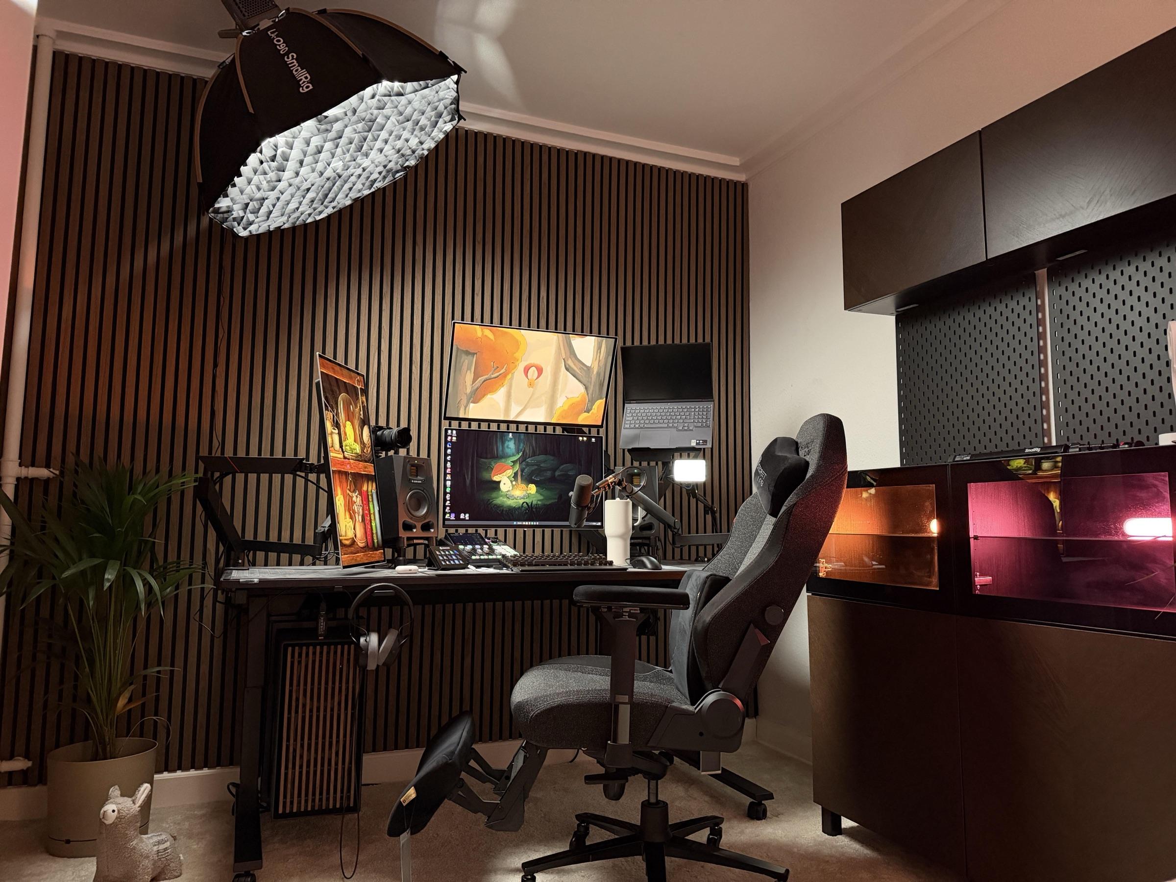 A dark room with a wood slat wall, three monitors on a black desk, a softbox overhead and lit display cabinets to the right.