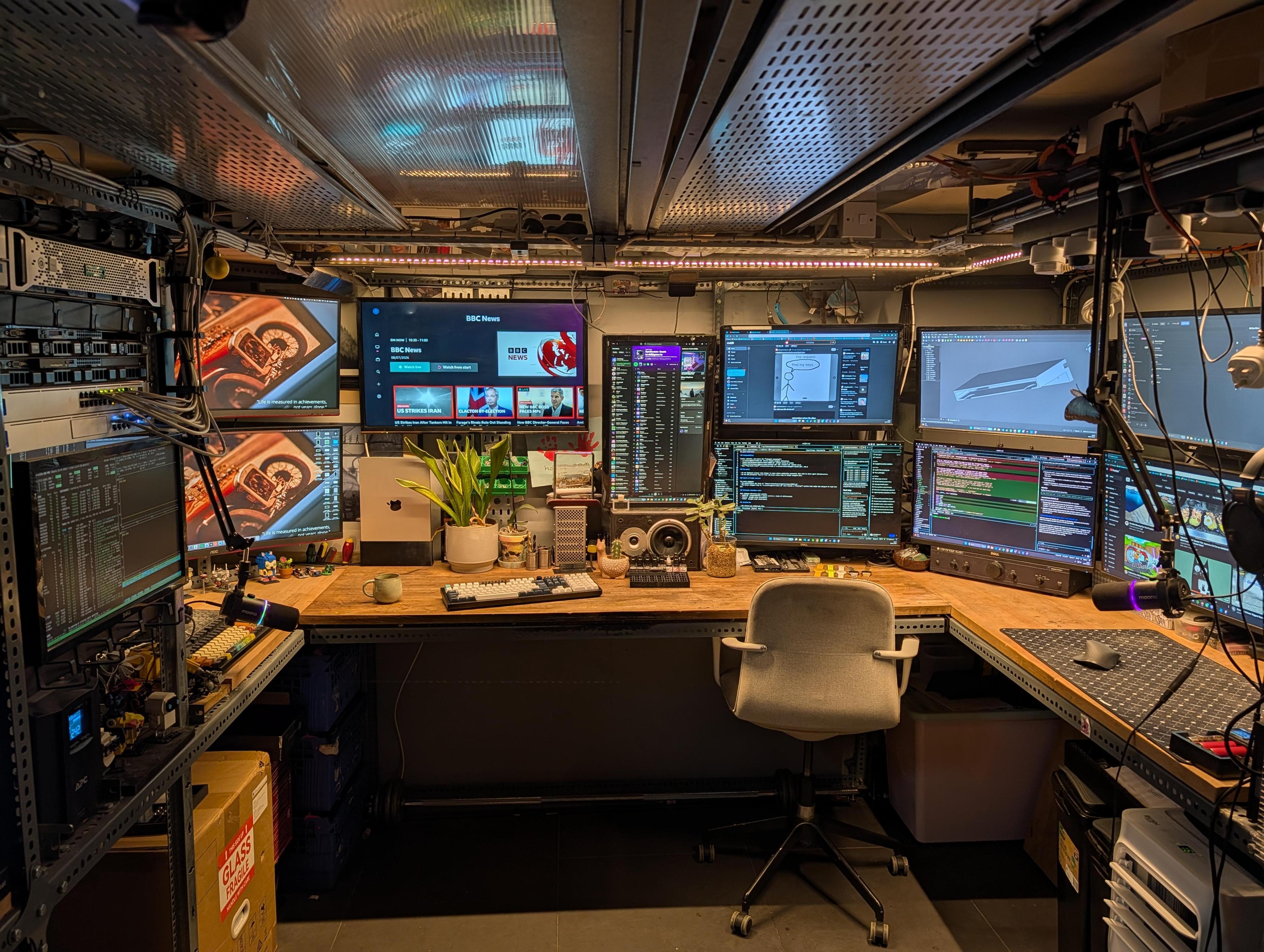 A low cellar office with a wooden worktop around three walls, a server rack on the left, and screens filling the wall above the desk.