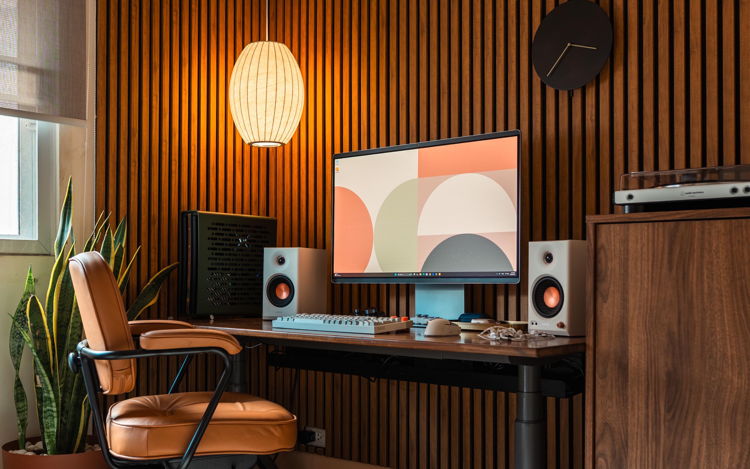 A warm wood slat wall behind a dark wood sit-stand desk, with one large screen, two cream speakers with copper drivers, a tan leather chair on a black frame, a paper pendant lamp, and a walnut cabinet with a turntable at the right.