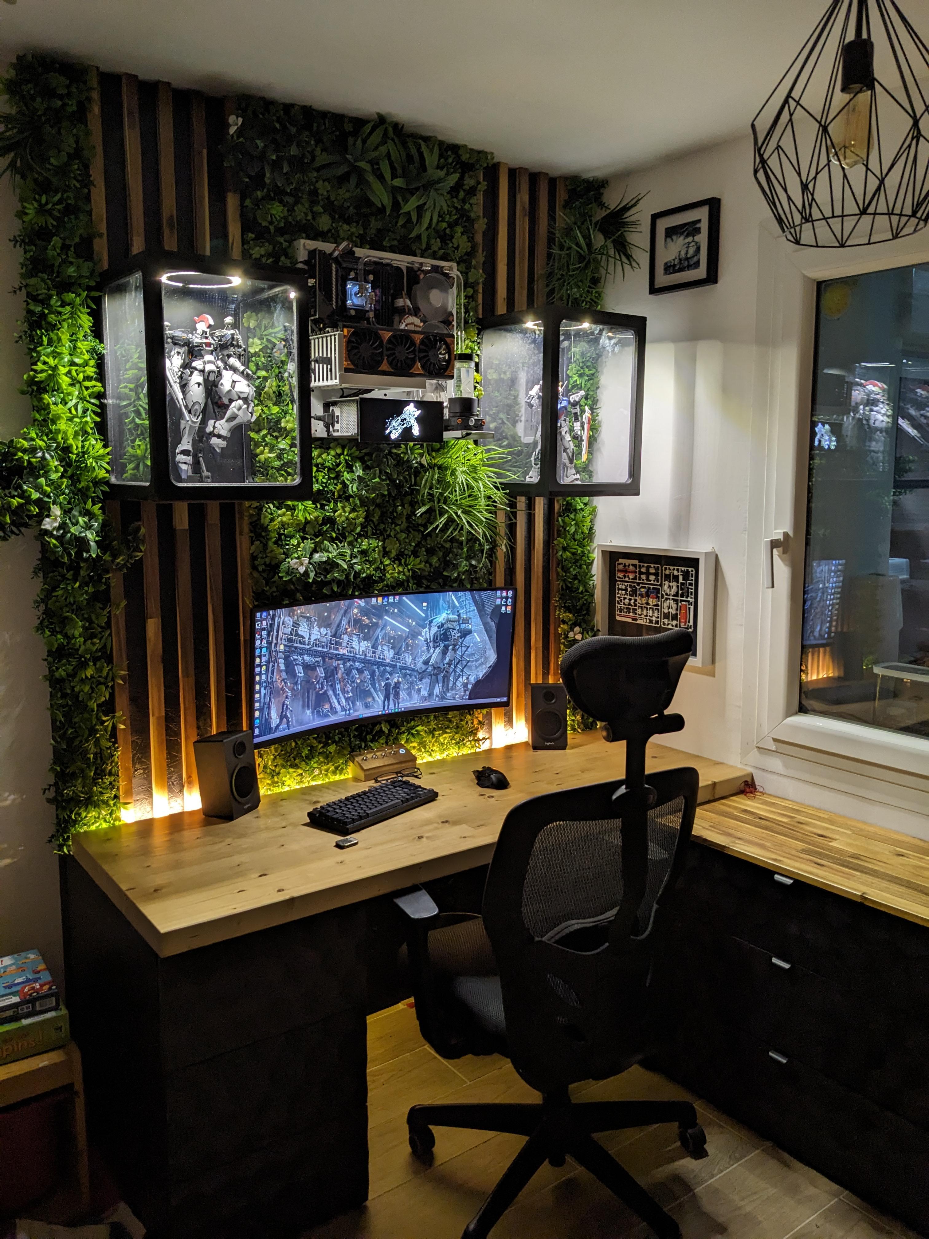 A corner desk with a pine top and an ultrawide screen, against a wall of artificial foliage over timber slats, with an open computer mounted on the wall between two lit cases of Gunpla.