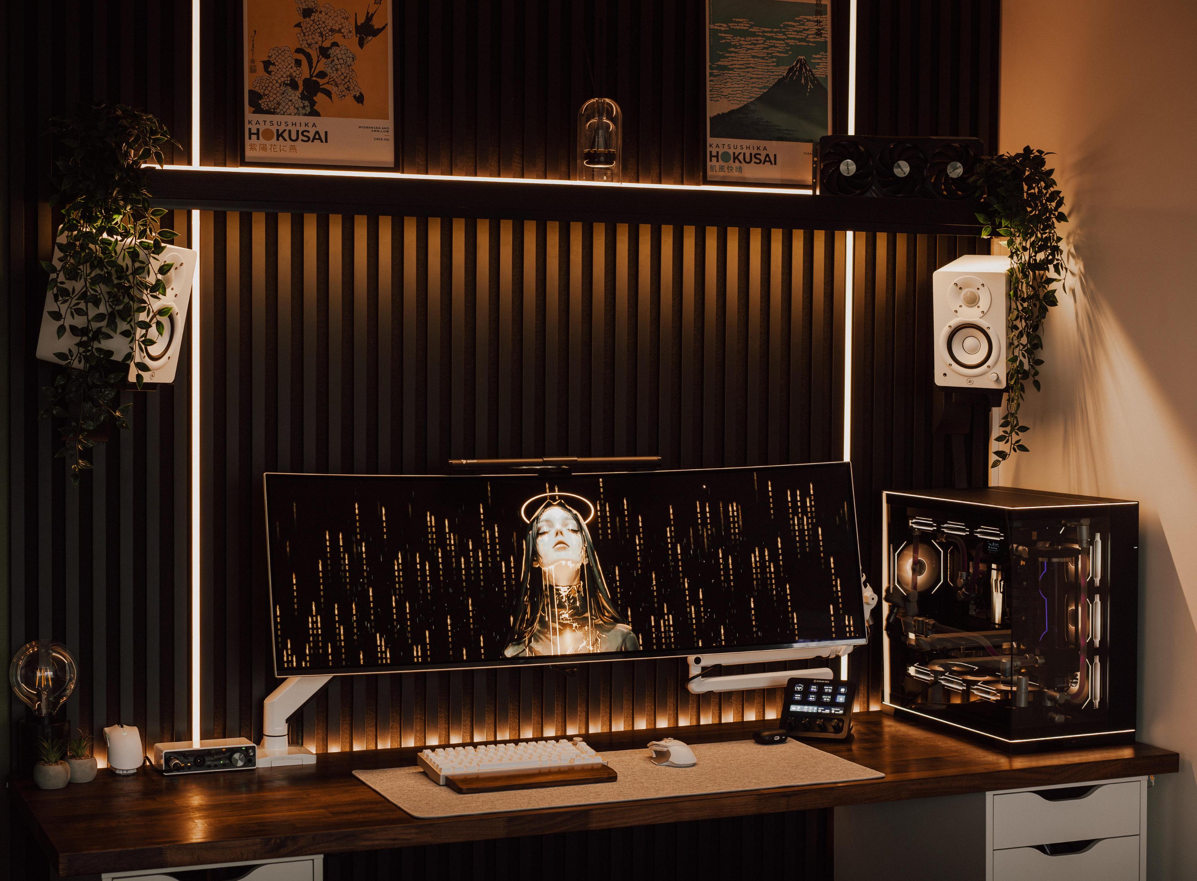 A dark room with a black vertical slat wall lit by white strips. A wide curved screen on a white arm stands on a walnut top over white drawer units, two white speakers hang on the wall either side, two Hokusai prints stand on a shelf above, and a water-cooled computer sits in a glass case on the right.