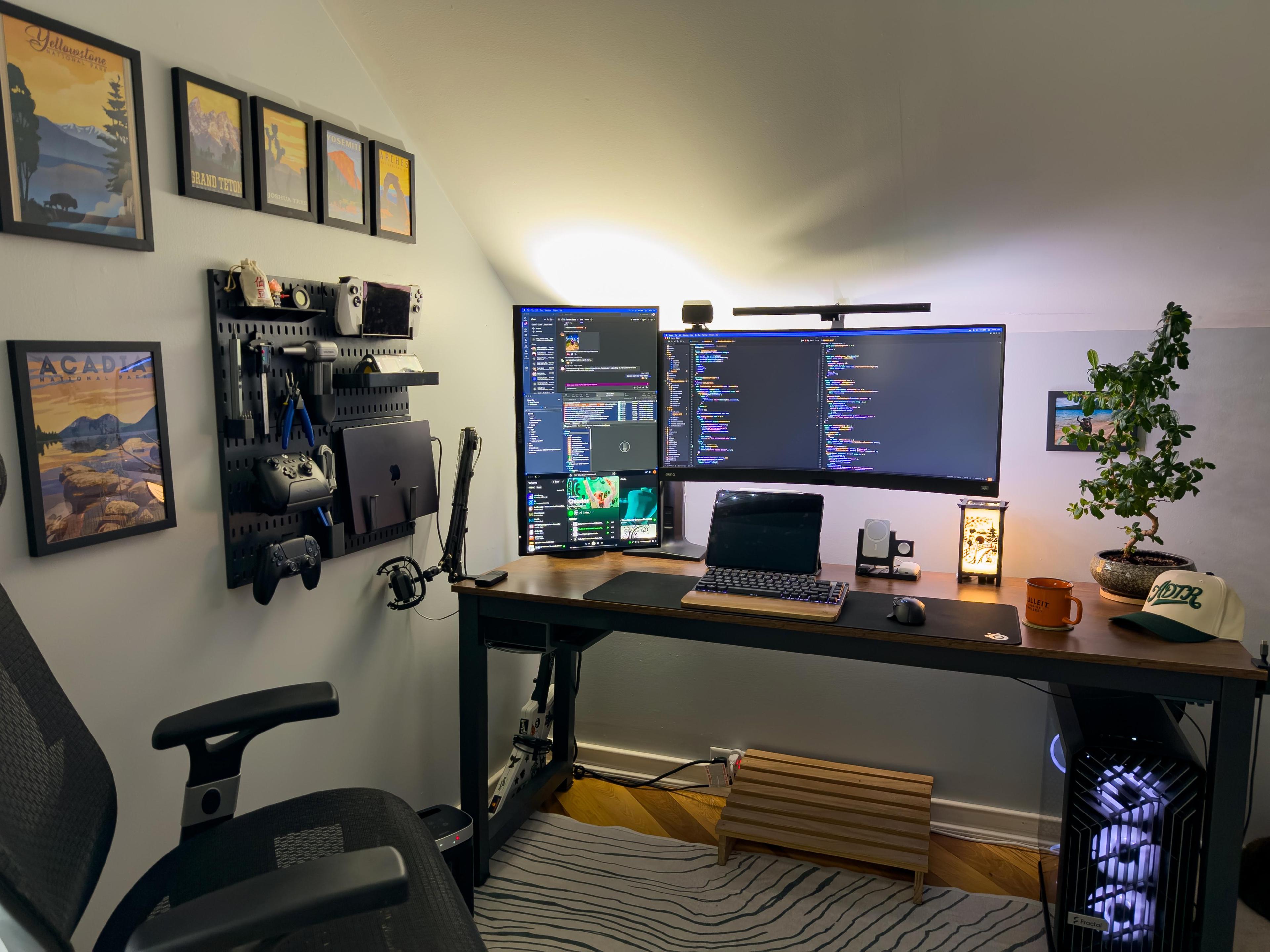 A desk in a corner under a sloping ceiling. Framed national park posters hang on the left wall above a black pegboard holding tools, controllers and a laptop. A curved ultrawide screen and a second screen stood on its end fill the desk, with a bonsai at the right end and a lit computer beneath.