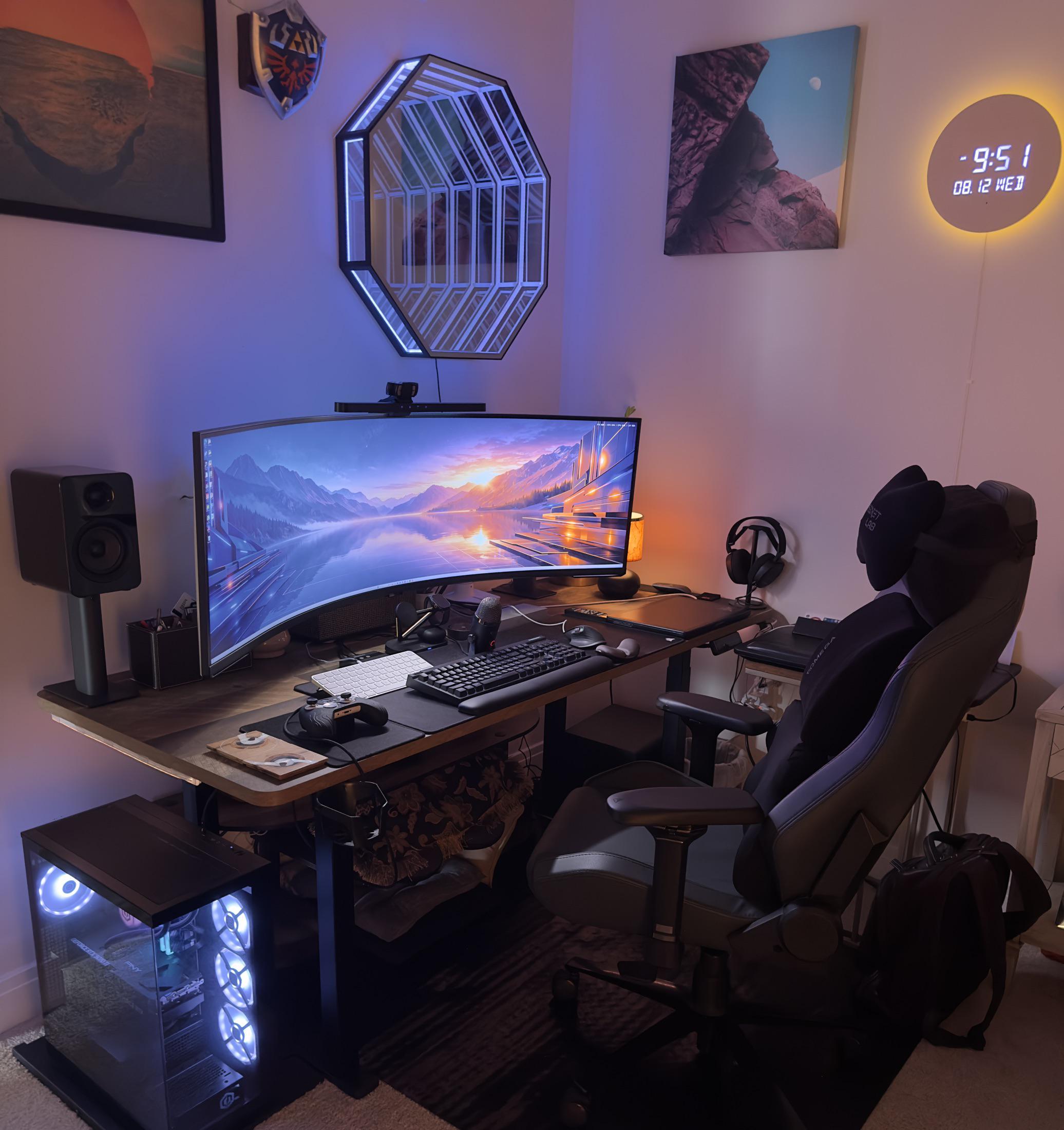 A corner desk lit blue and purple, with one very wide curved monitor, an octagonal lit mirror on the wall above it, a round orange clock to the right, a black speaker on a pillar and a PC glowing on the floor.