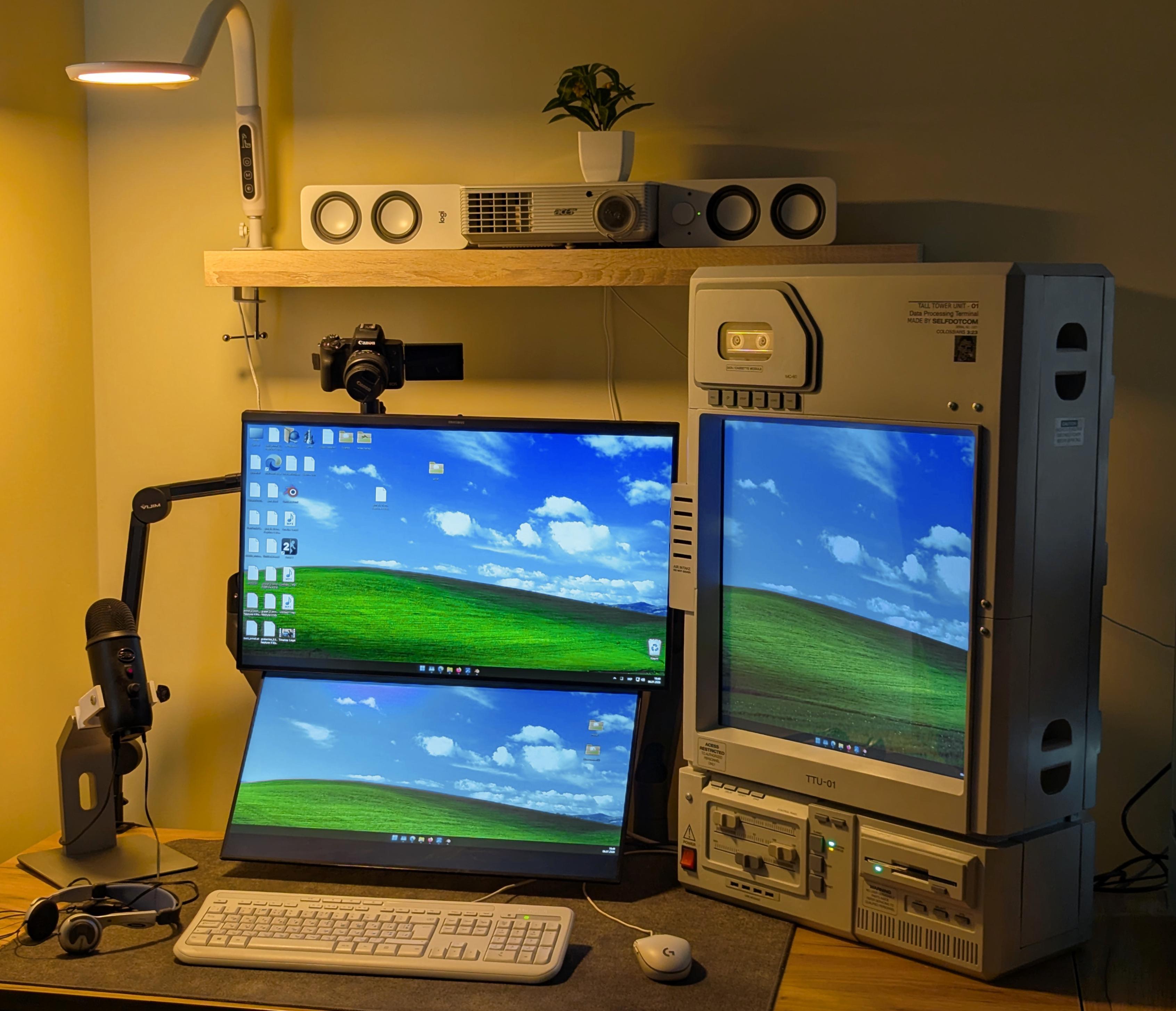 A warm lit corner desk with a tall hand built white terminal on the right, two screens showing a green hill under a blue sky, and a white keyboard.
