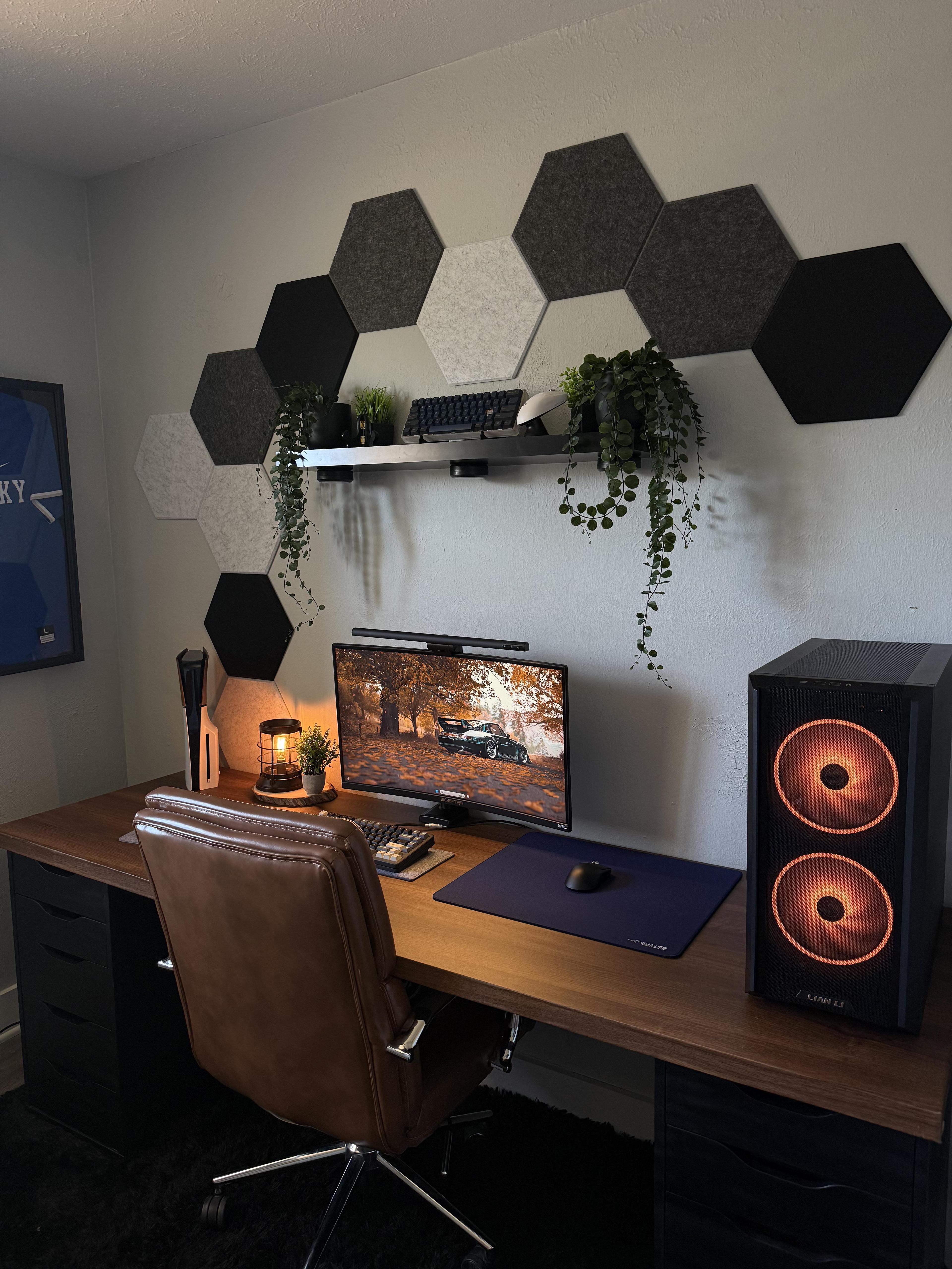A warm wood desk top on two black drawer units, with a curved screen under a light bar, a brown leather chair, a wall of grey and black hexagon felt panels above, and a black tower with orange lit fans at the right end.