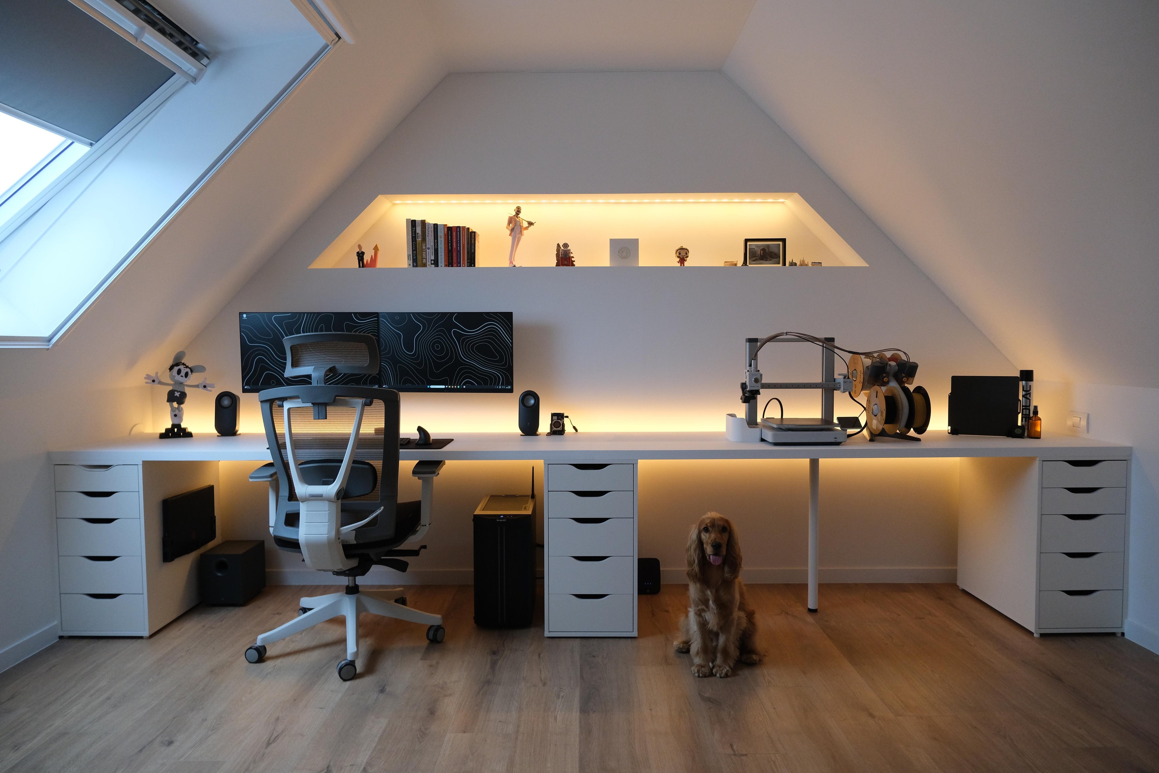 A white attic room under a pitched roof, with a long white desk along the whole wall, a lit recess shelf of books above it, two screens on the left, a 3D printer on the right, white drawer units under the desk and a golden dog sitting on the wooden floor.
