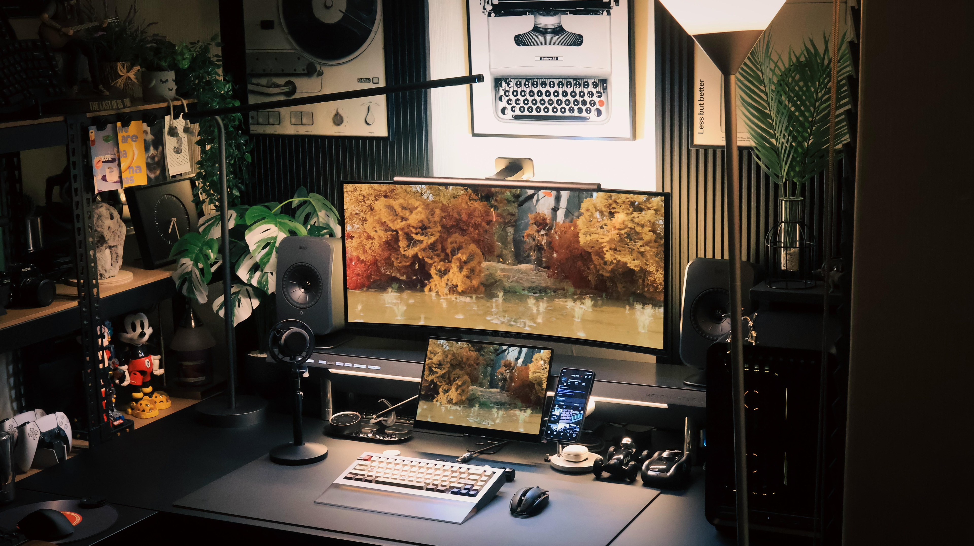 A dark desk lit warm, with a curved ultrawide screen, a small portable monitor propped in front of it, a microphone on a stand, black industrial shelving of plants and figures to the left, and a black PC tower to the right.