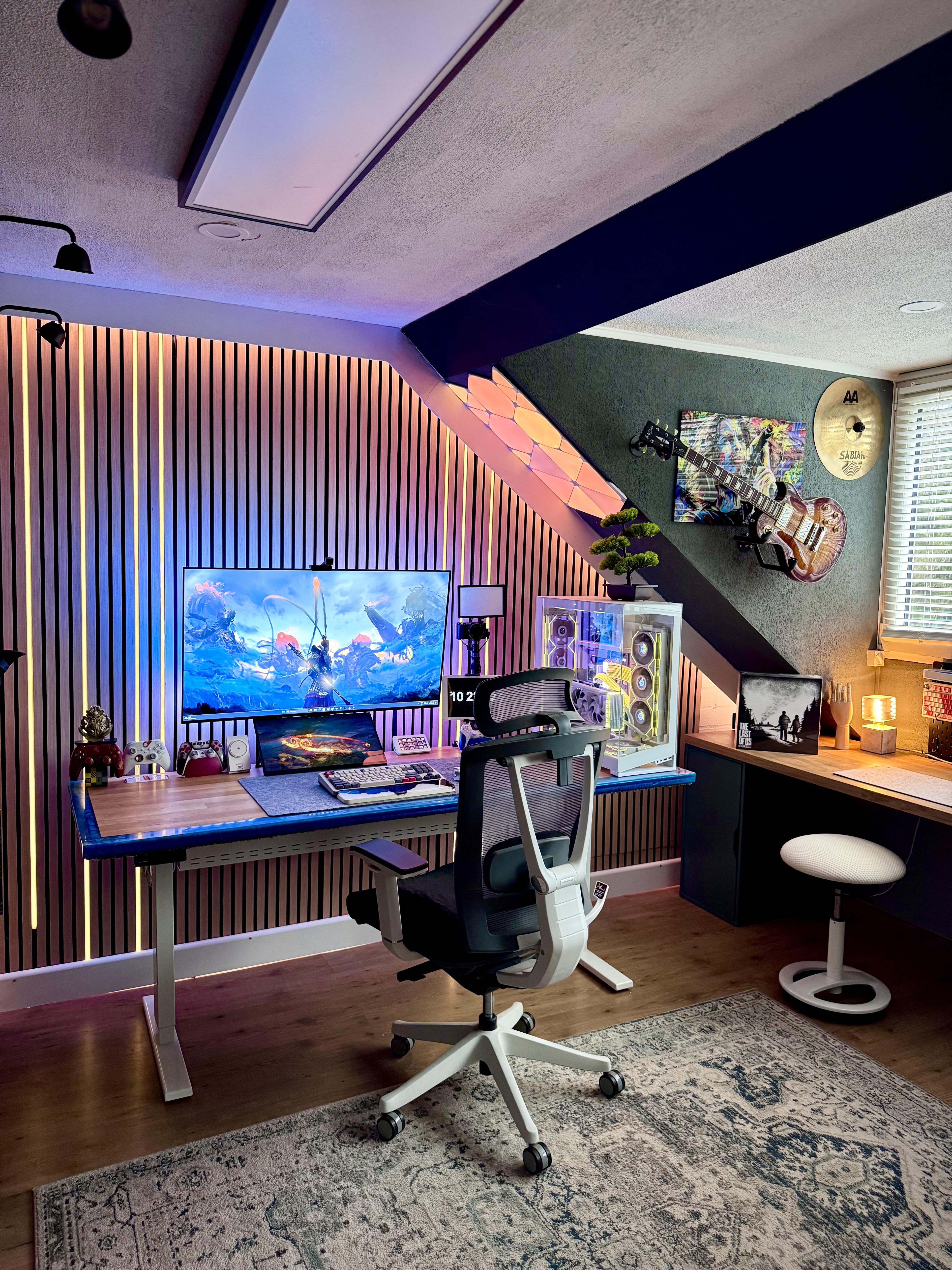 A desk in an attic corner under a sloped roof, against a wall of wood slats lit from between, with a large screen, a small screen under it, a white PC case and a white mesh chair on a rug.