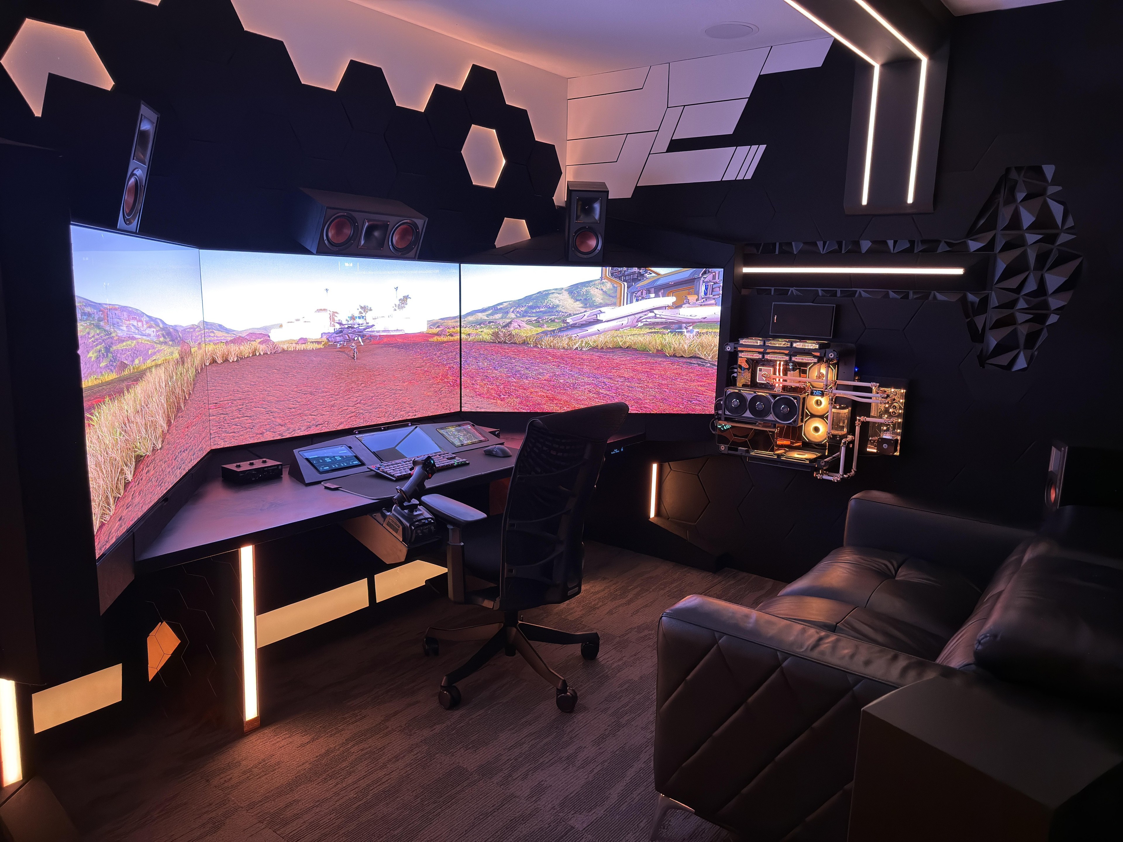 A black basement room with three curved screens in an arc, hexagon panels on the walls, a plywood desk with a flight stick, and an open water-cooled computer mounted on the right-hand wall.