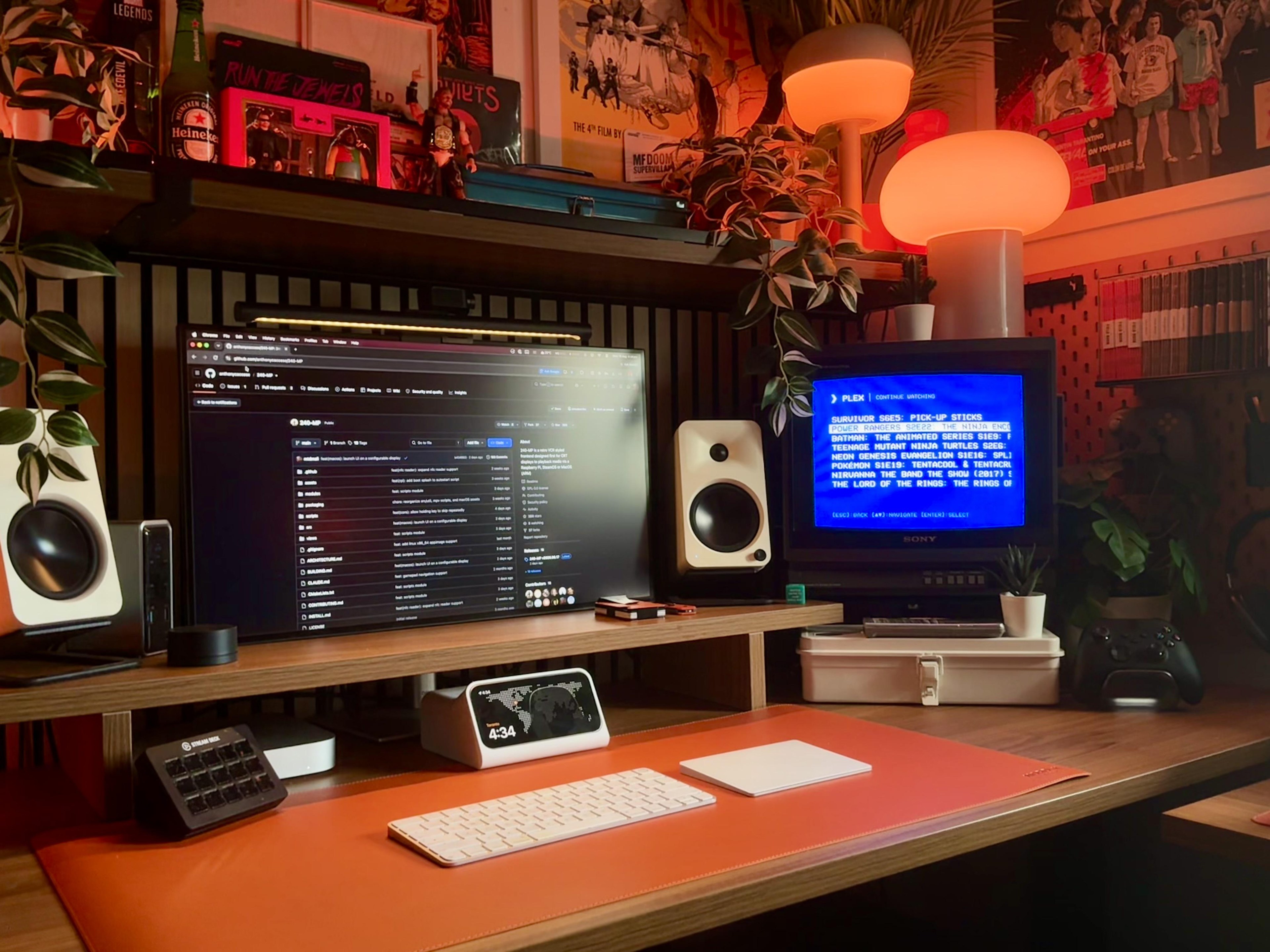 A warm lit desk with a wide monitor on a wooden shelf, a small Sony CRT beside it showing a blue Plex menu, cream speakers at each end, and a shelf of posters, figures and trailing plants above.