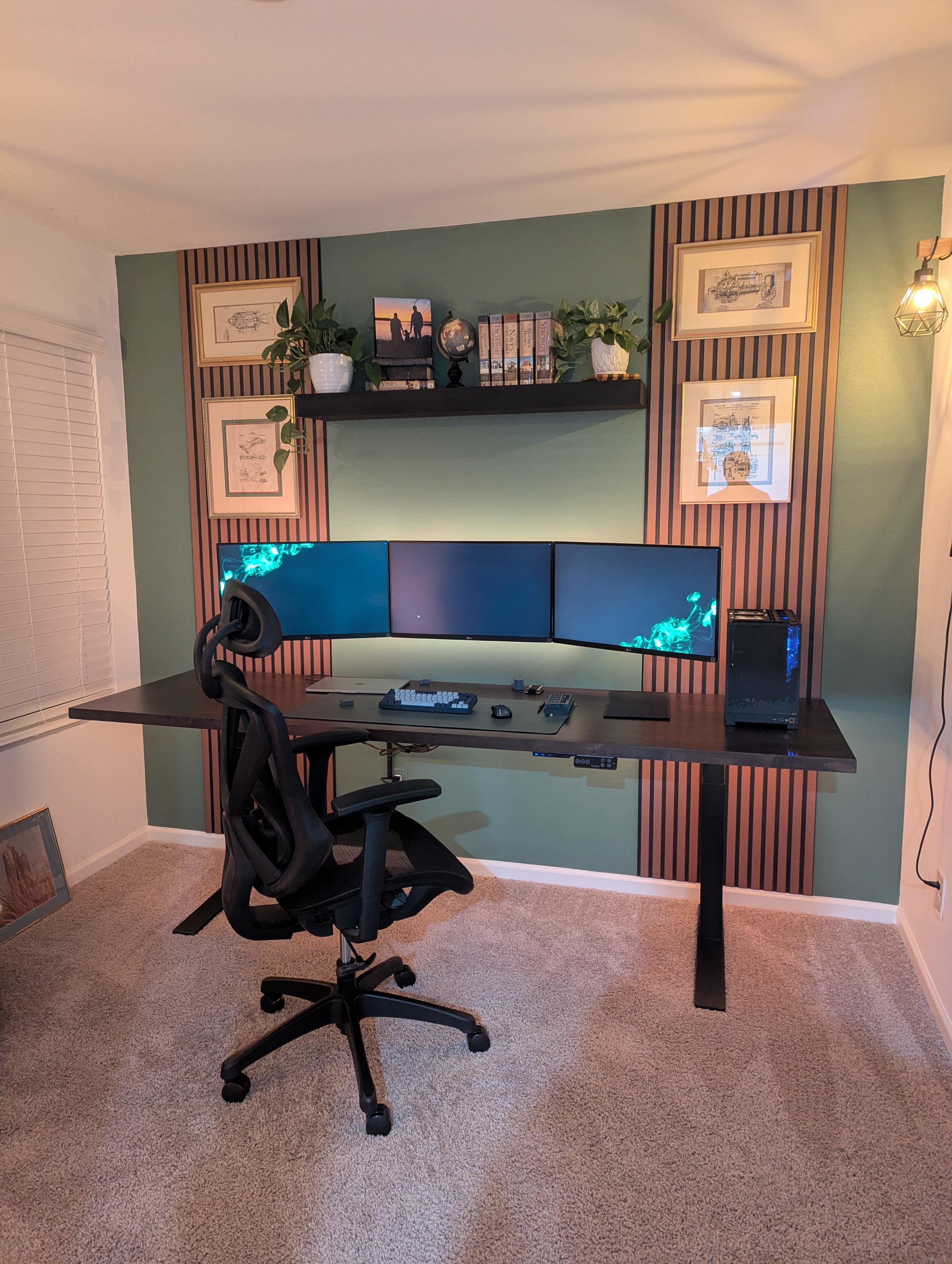 A green wall with vertical black and wood slat panels, three monitors that appear to float on it, a long dark sit-stand desk with a keyboard and a PC tower, and a black mesh chair on beige carpet.