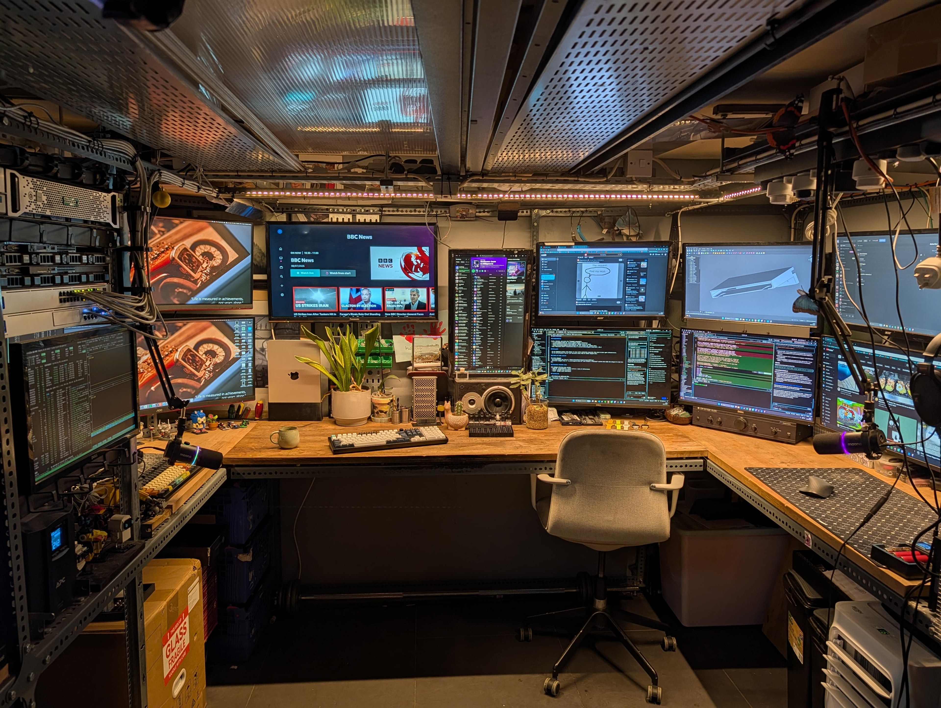 A low cellar office with a wooden worktop around three walls, a server rack on the left, and screens filling the wall above the desk.