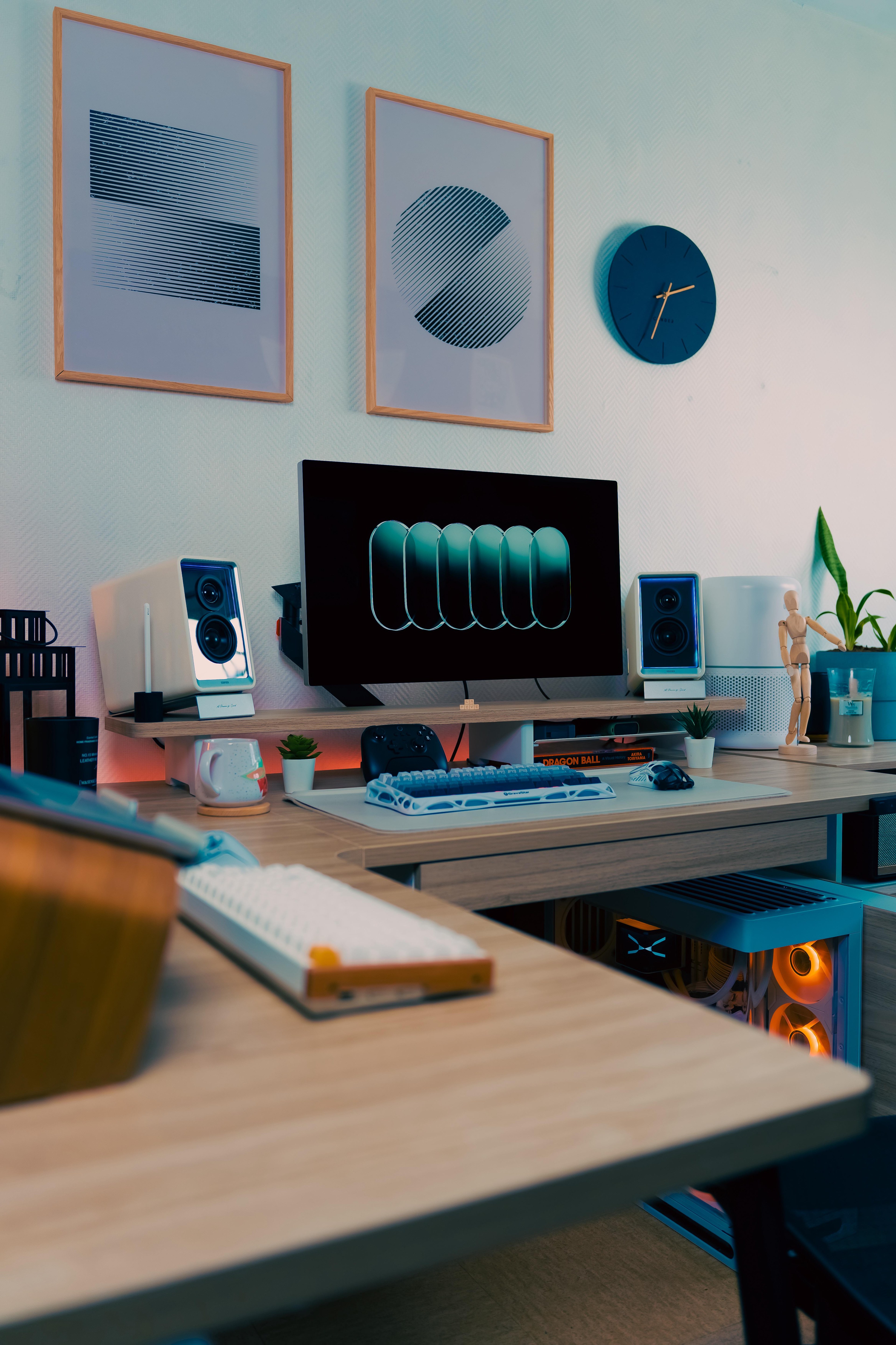 An L shaped desk in pale wood against a white wall, with one dark screen on a wooden riser, white speakers either side, two keyboards, and a pale blue PC glowing orange in the shelf below.