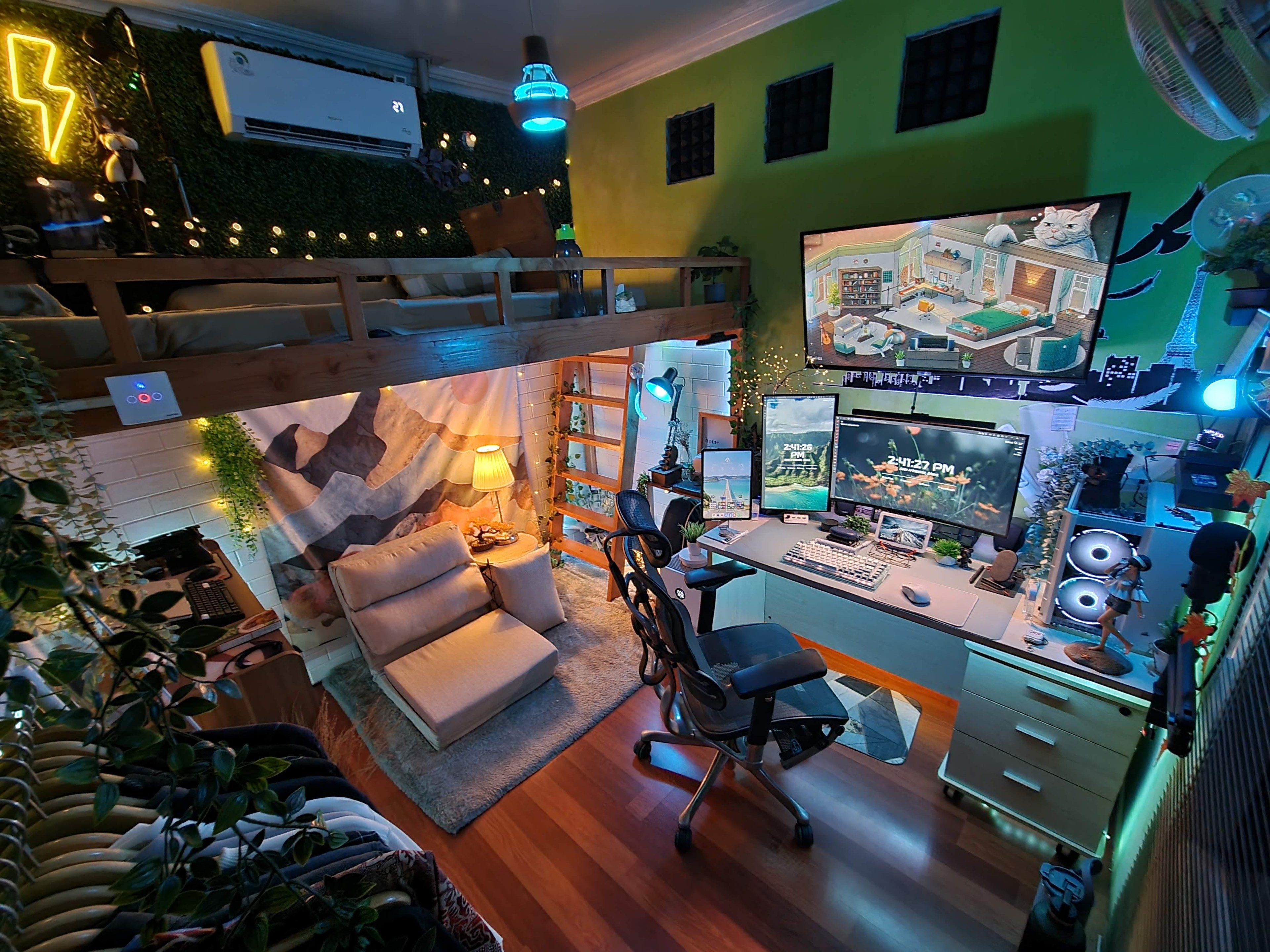 A small green room shot from above, with a timber loft bed over a low sofa and a mountain mural, artificial grass and string lights along the loft, and a corner desk with two screens, a tablet and a mesh chair.