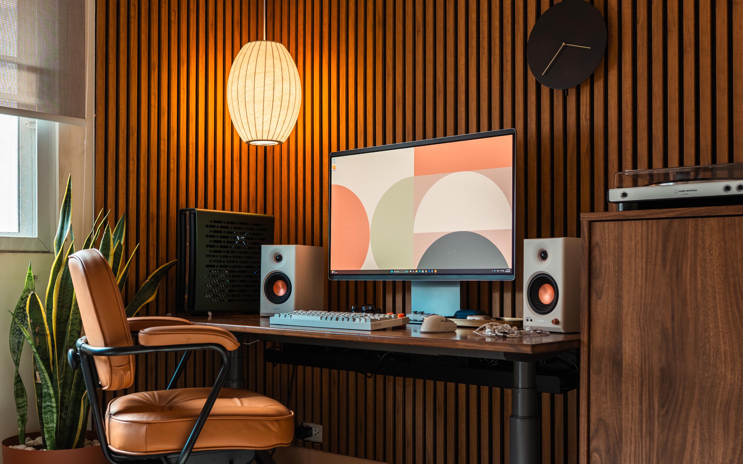 A warm wood slat wall behind a dark wood sit-stand desk, with one large screen, two cream speakers with copper drivers, a tan leather chair on a black frame, a paper pendant lamp, and a walnut cabinet with a turntable at the right.