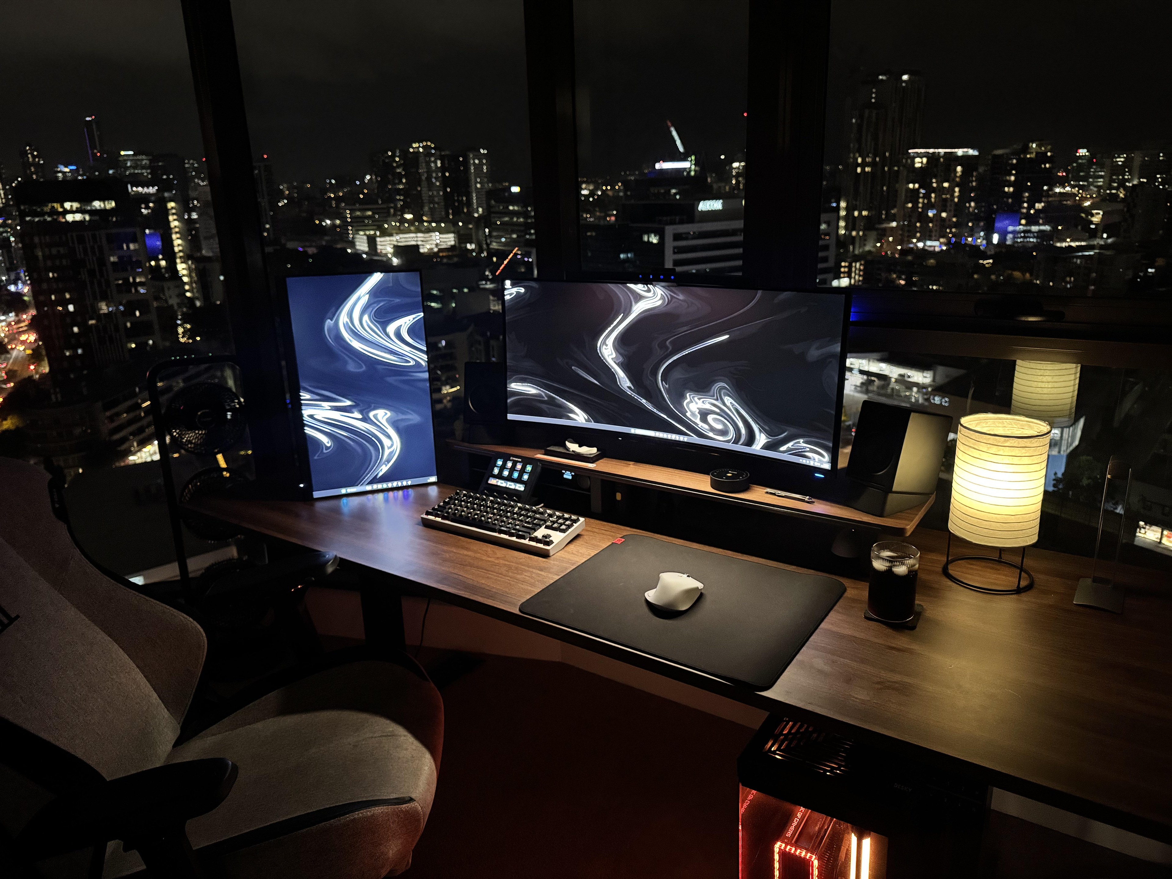 A wooden desk in the corner of a high floor flat at night, with floor to ceiling glass on two sides and the lights of Brisbane behind it, a curved main screen on a wooden shelf, a second screen stood on end to the left, a mechanical keyboard, a white mouse on a black mat and a lit paper lantern lamp on the right.