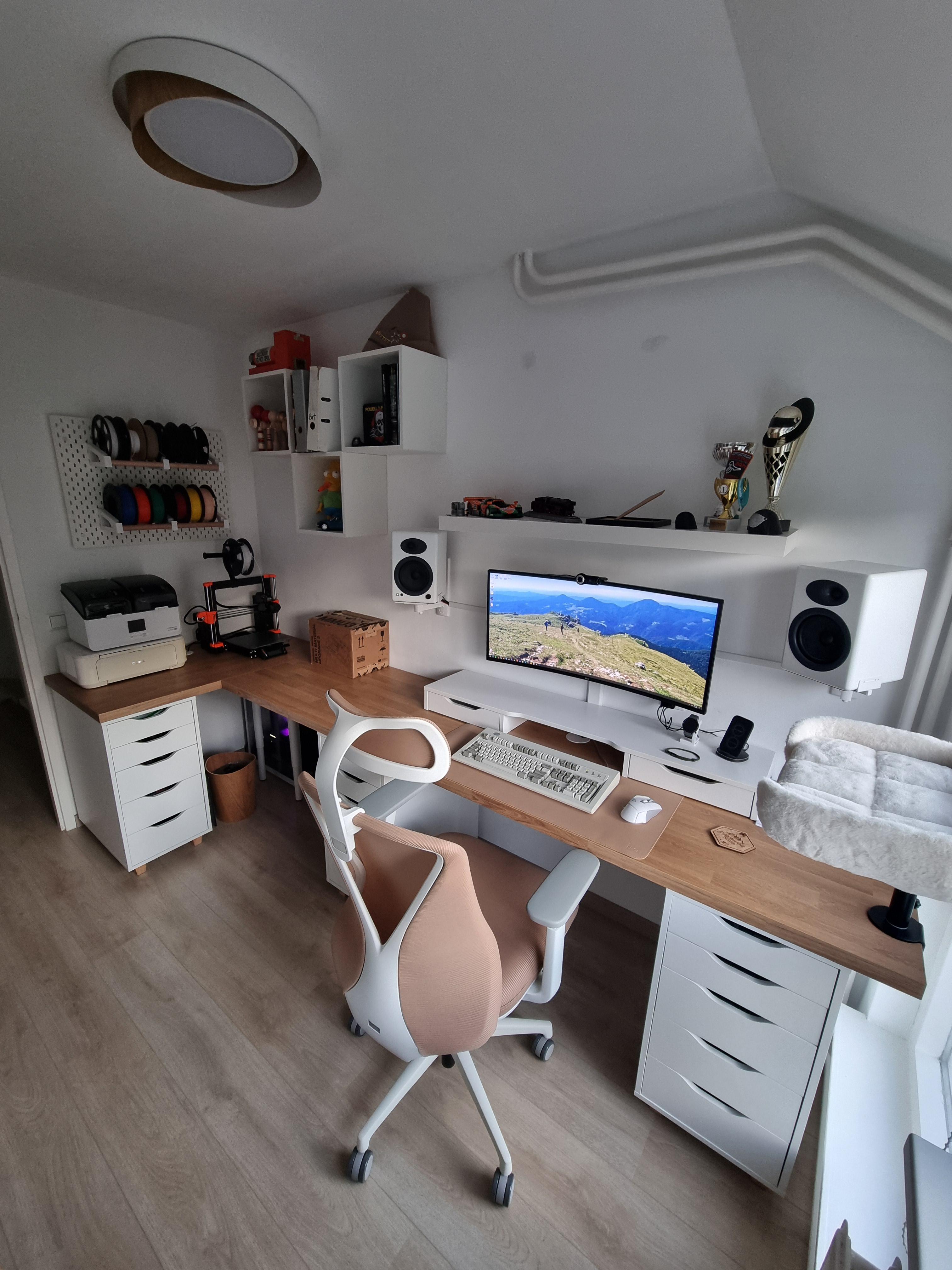 A white attic room with a long wooden desk running wall to wall, an ultrawide monitor on a riser, white speakers on wall mounts either side, a white and pink chair, and a 3D printer with a rack of filament at the left end.