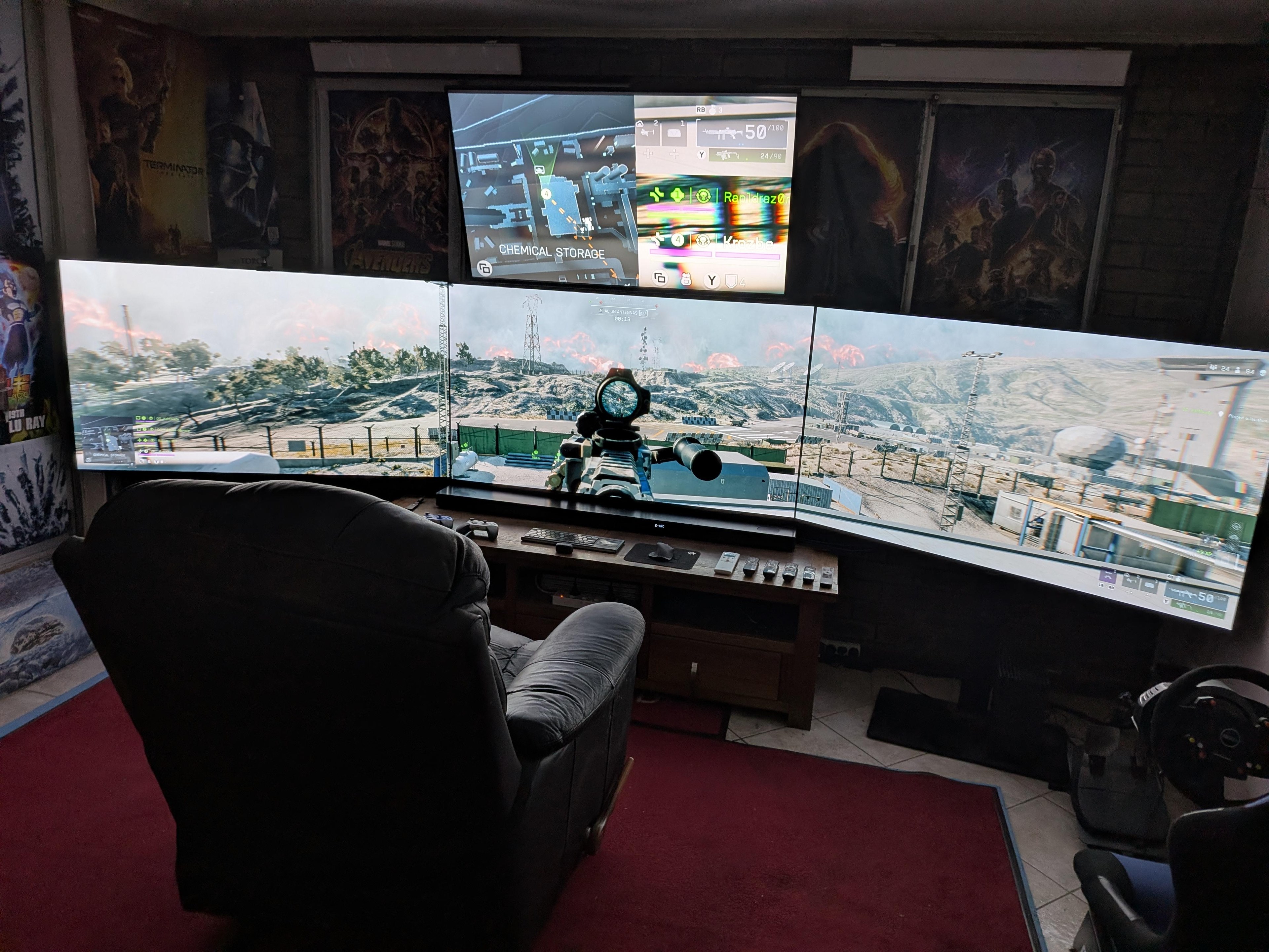 A dark living room with three big screens in an arc, a fourth screen above them, a black leather recliner in the middle and film posters on the walls.