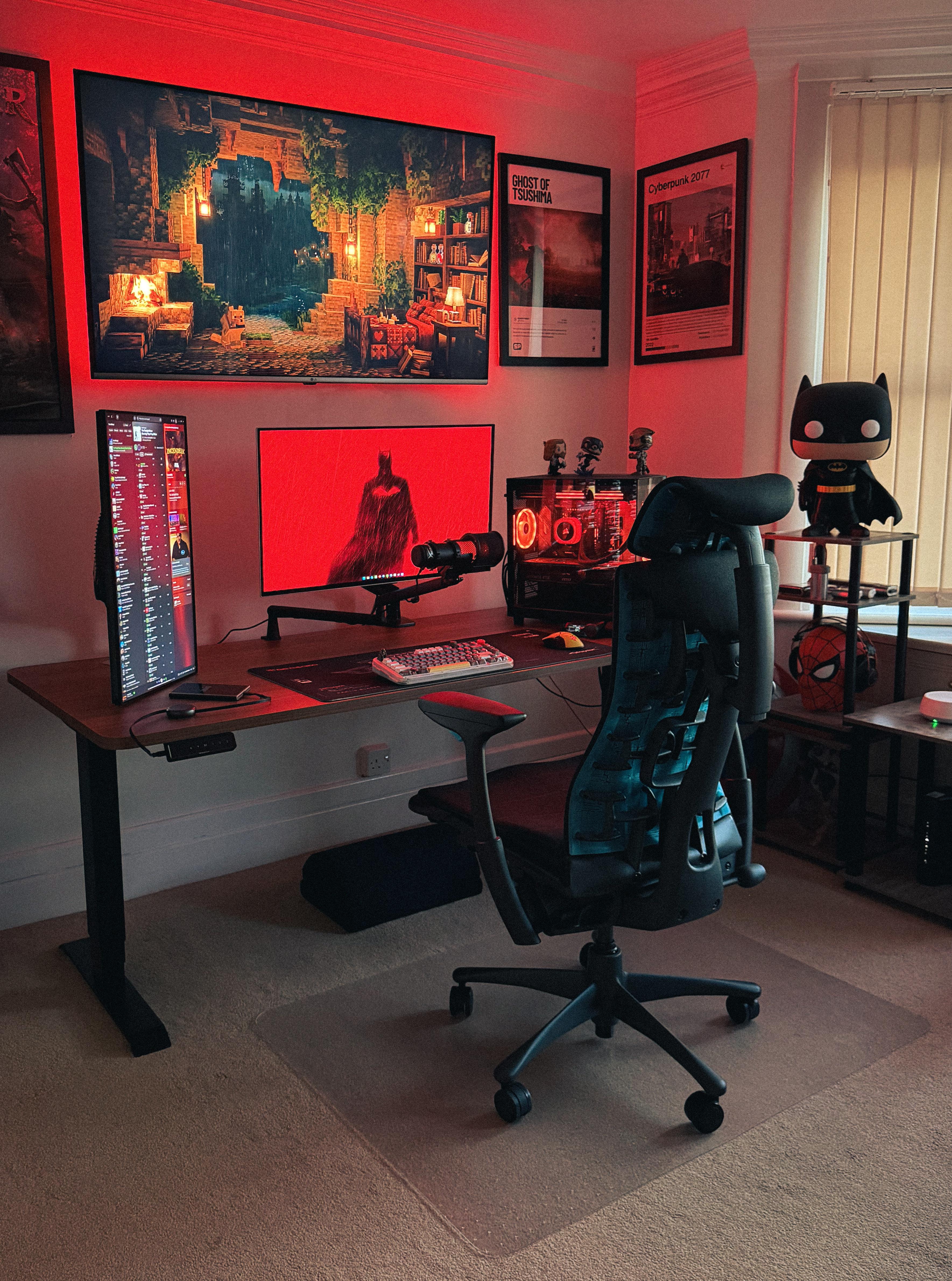 A red-lit bedroom corner with a wall-mounted TV showing a Minecraft cave, a wide monitor on a walnut desk, a second monitor stood on end, a glass PC with a red water loop, a black mesh chair, and a large Batman figure on a shelf.