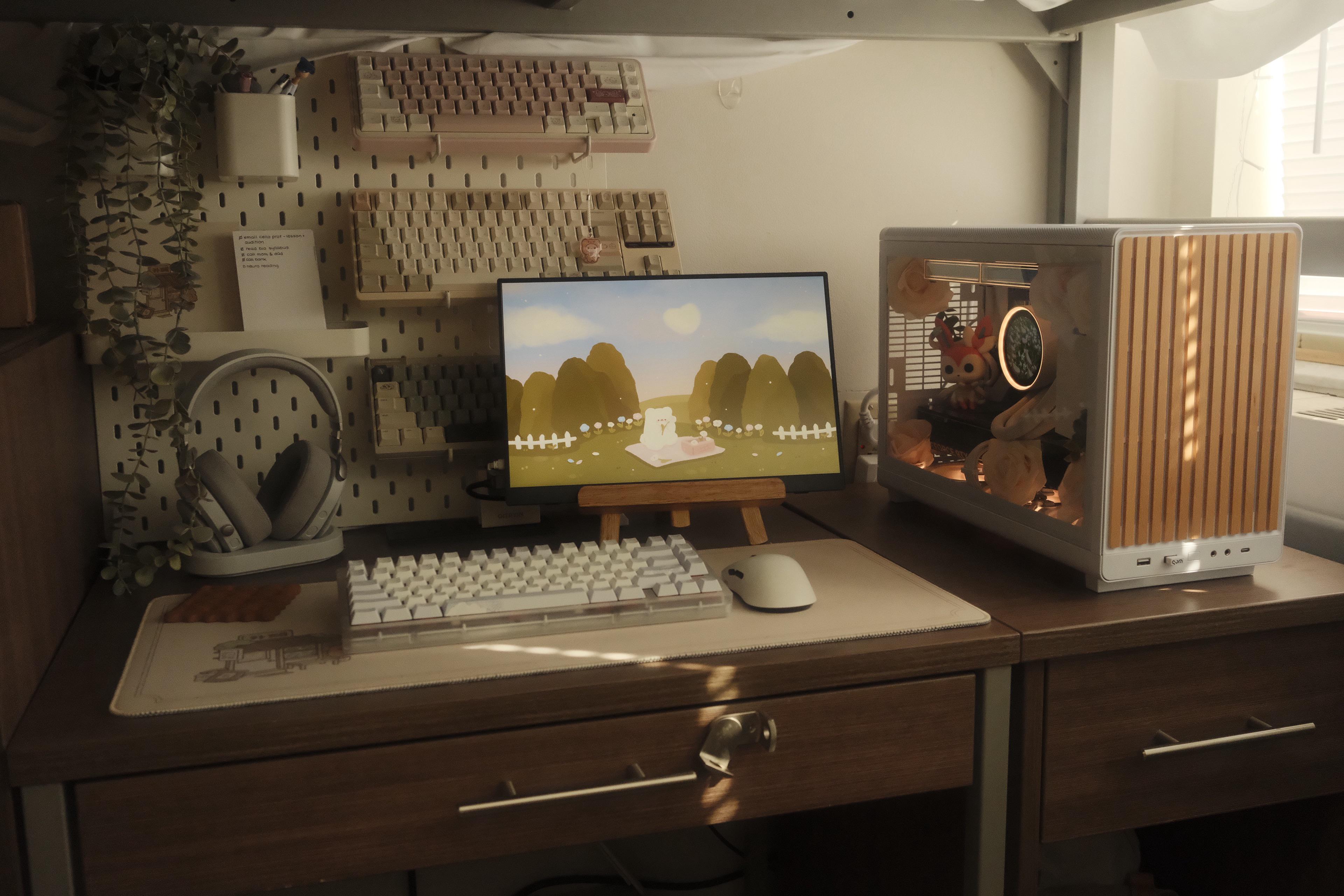 A dorm desk in warm afternoon light, with a small screen on a wooden easel, a cream pegboard of keyboards and a headphone stand behind it, a white keyboard and mouse on the desk, and a small white PC with dried flowers inside.