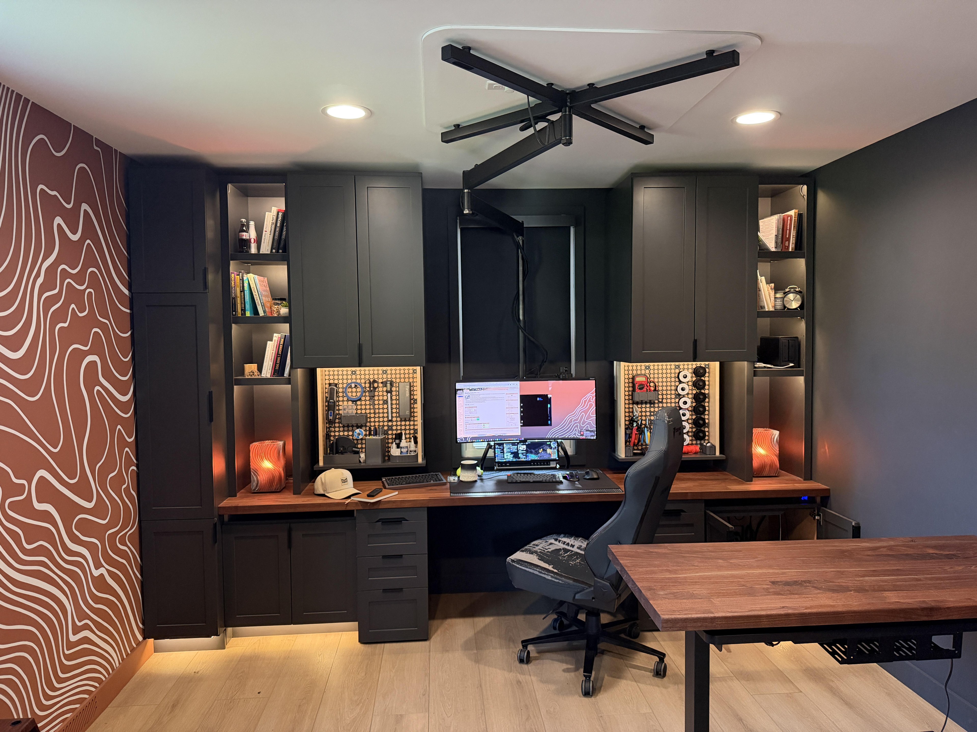 A home office with a dark built-in wall of cabinets and open shelves over a walnut worktop, a lit pegboard at each end, and an ultrawide screen hanging from a jointed steel arm fixed to the ceiling. A rust topographic mural covers the wall on the left.