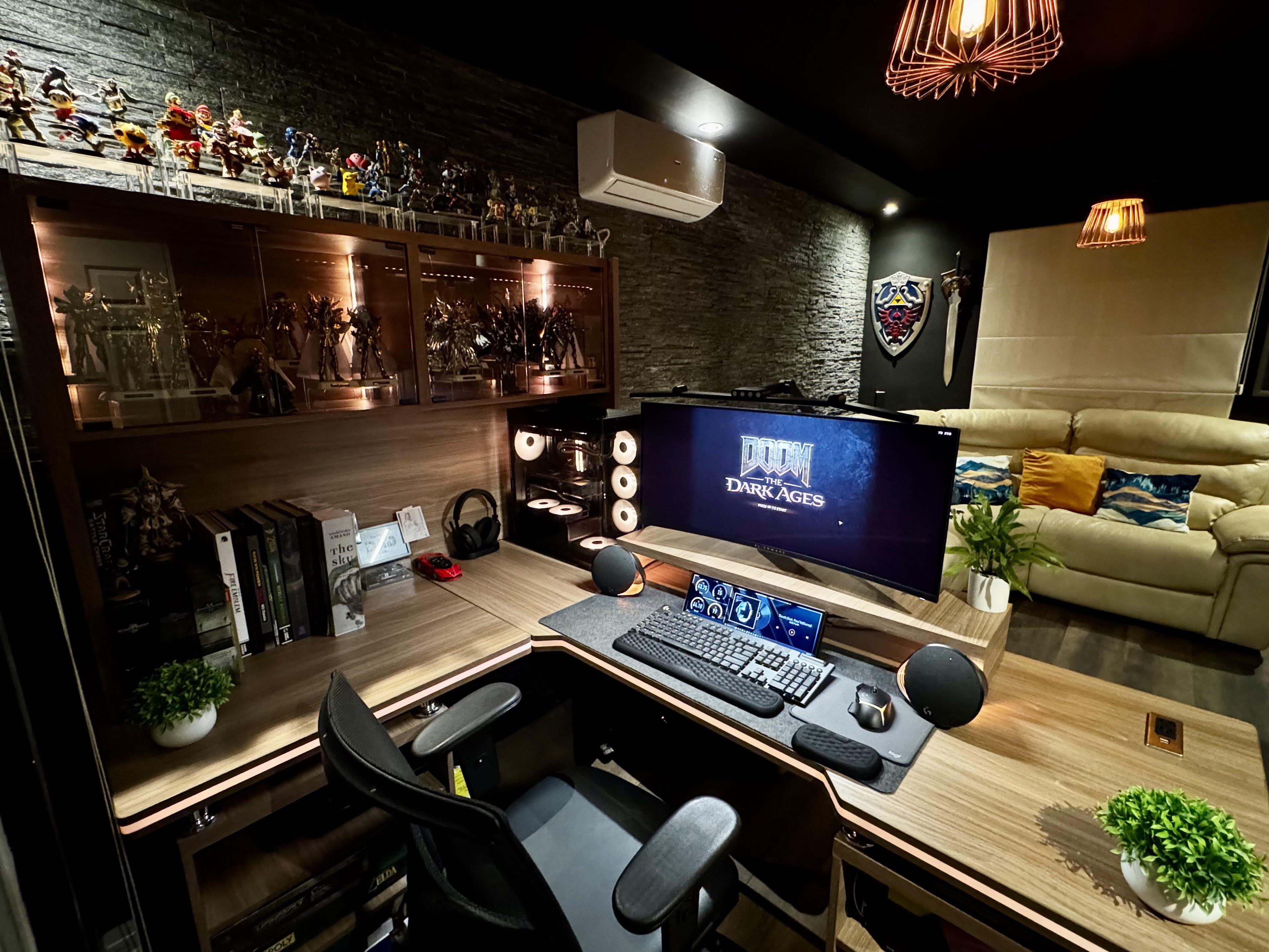 A dark corner room with a stone wall and wood lining. A lit glass cabinet of gold armoured figures stands over an L-shaped wooden desk holding a curved screen, a keyboard and a wide strip display. A shield and a sword hang on the wall, and a cream sofa fills the right of the room.