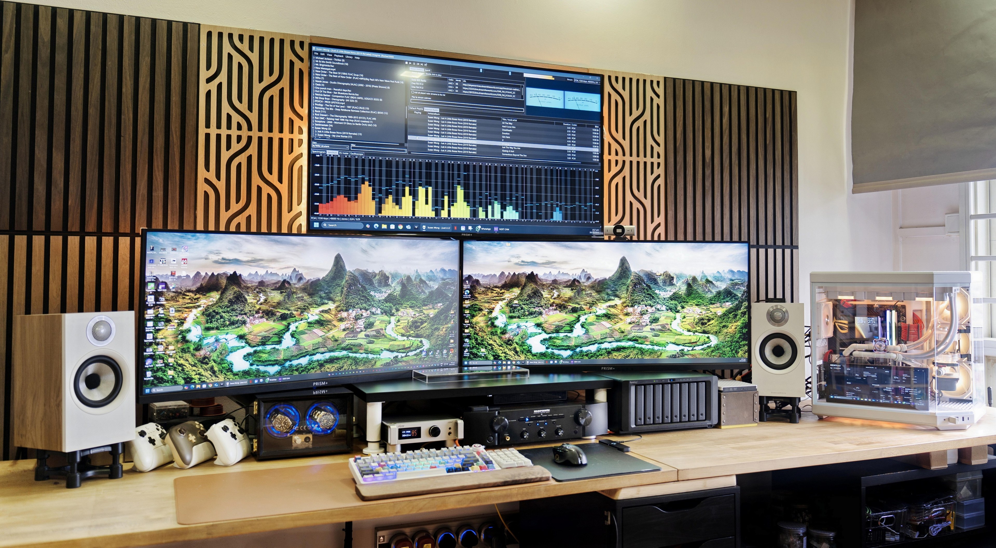A wide light wood desk against a dark slatted wall, with two ultrawide screens below a third screen, white bookshelf speakers on stands at both ends, a hi-fi amplifier and a network drive under the middle, and a white water-cooled computer in a glass case on the right.