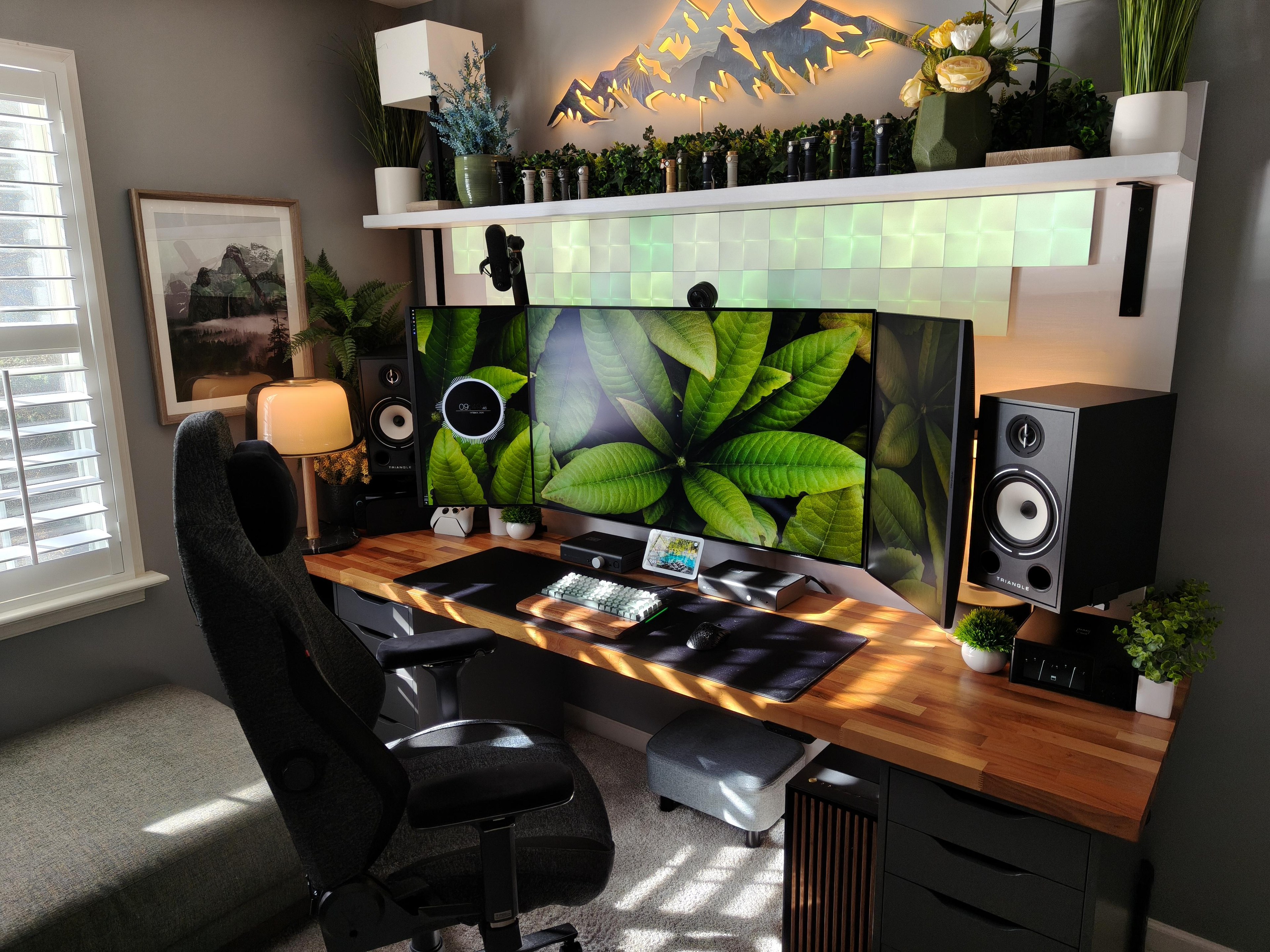 A desk in morning sun. A wide screen sits between two screens stood on their ends, all mounted on a white panel built out from the wall. A walnut butcher block top rests on black drawer units, with a tall speaker at each end and a shelf of plants, flashlights and a lit metal mountain range above.