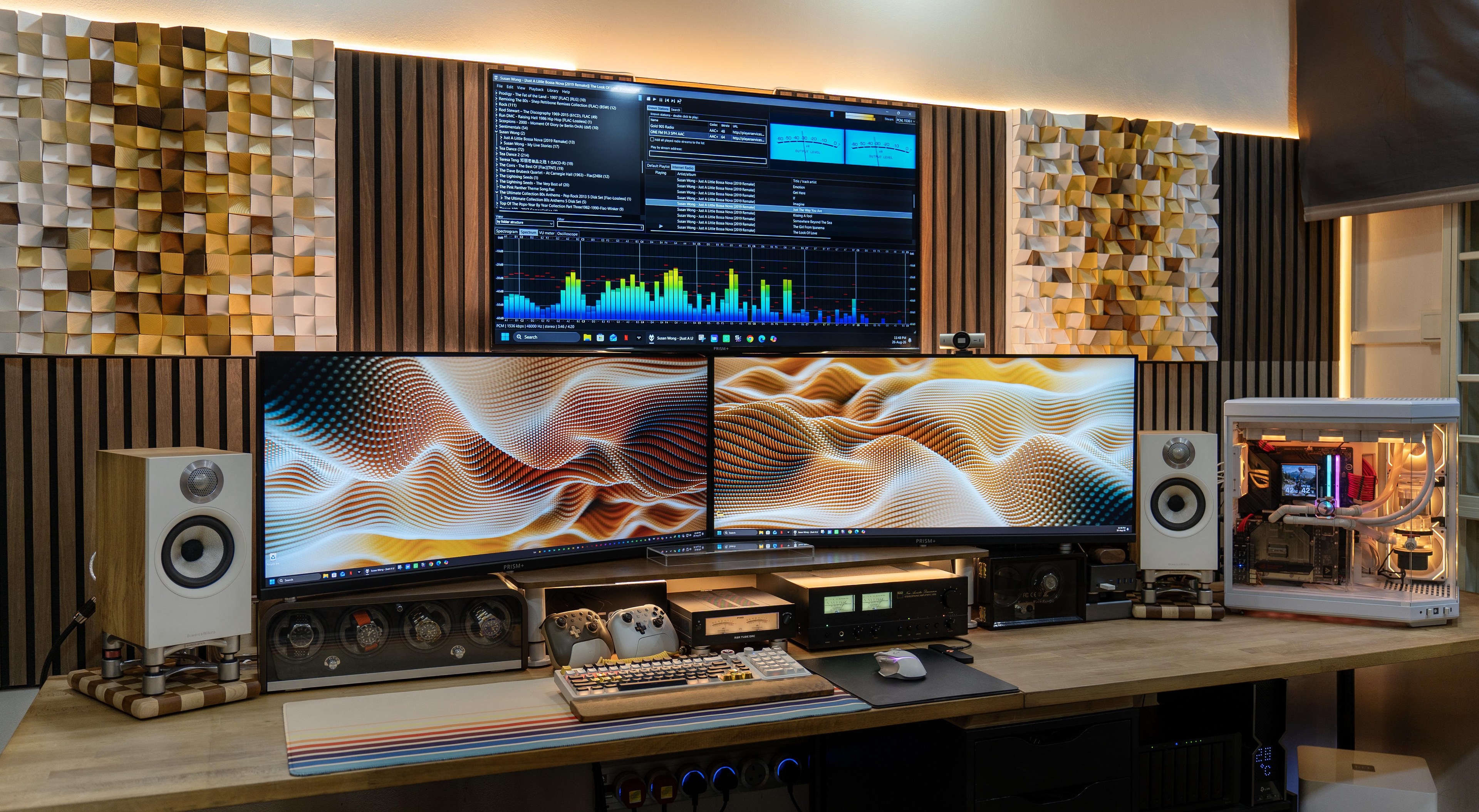 A wide wooden desk against a wall of wood slats, with two large monitors side by side under a TV, a white bookshelf speaker at each end on a stand, a row of audio boxes under the screens, and a white water cooled PC on the right.